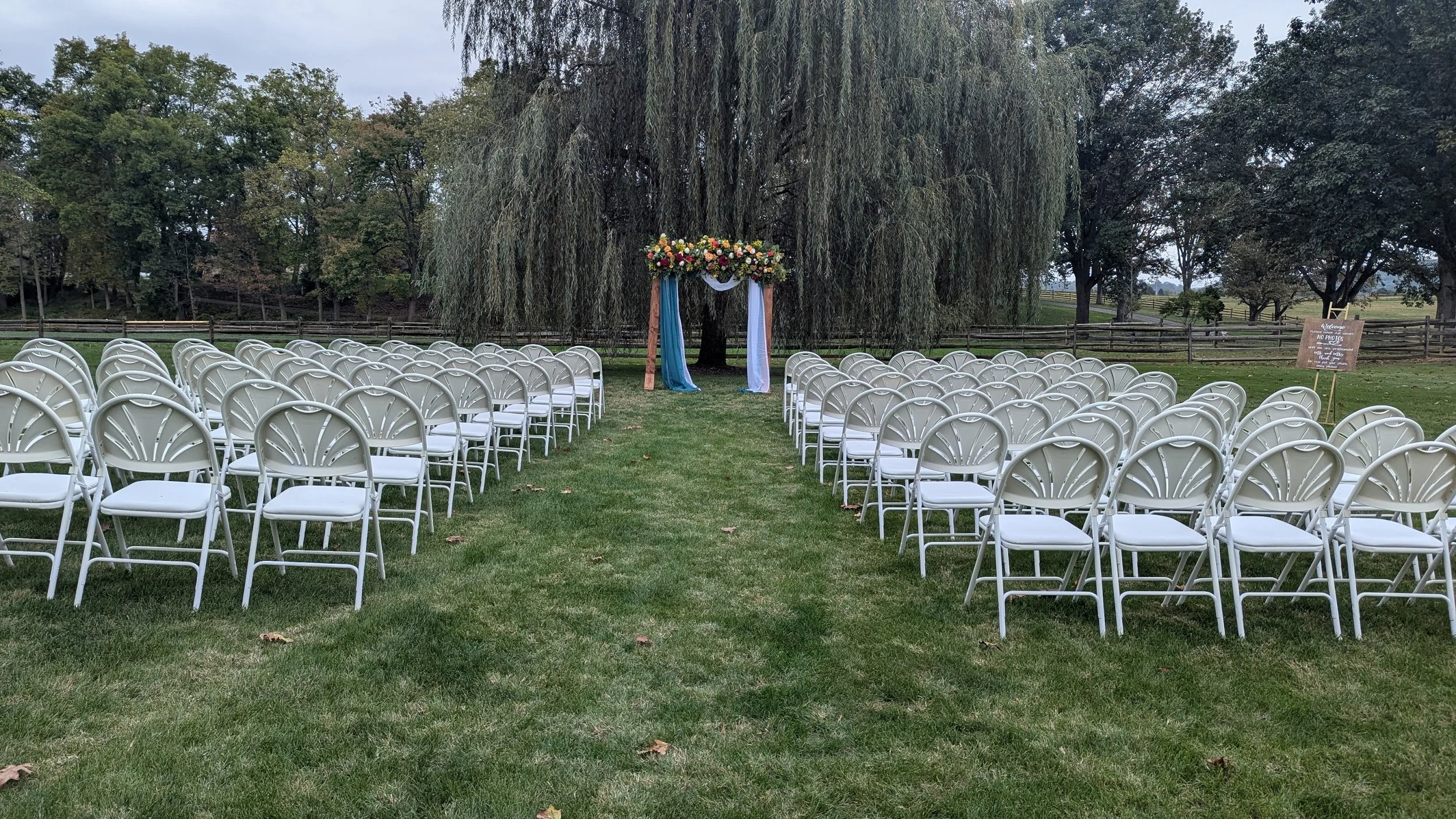 An outdoor wedding setup with white chairs arranged in rows on a grassy lawn, facing a decorated arch under a large weeping willow tree, with trees and a wooden fence in the background.