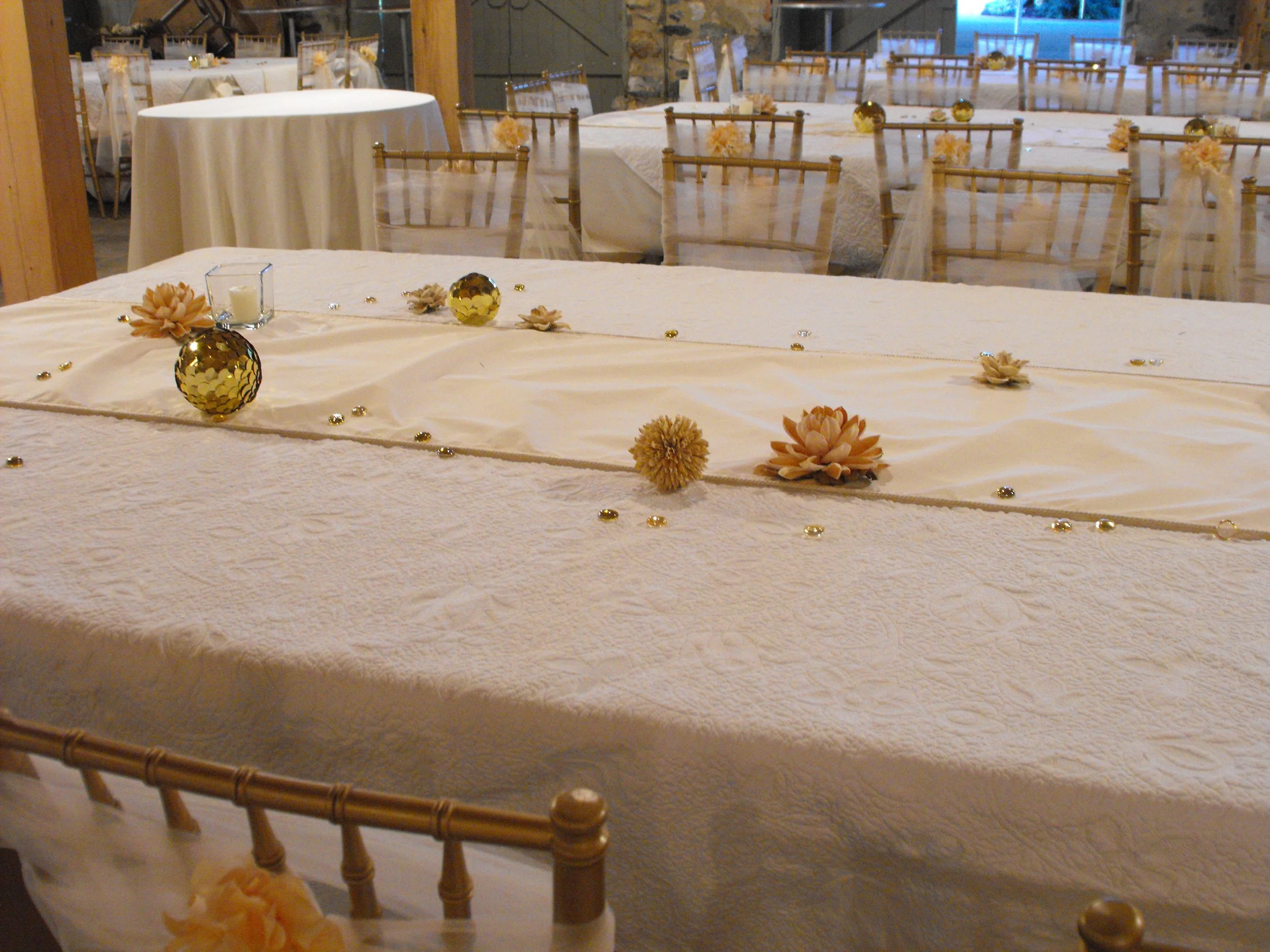 Decorated banquet table with gold and peach floral accents, candles, and scattered gemstones, set in a reception hall with multiple round tables and chairs.