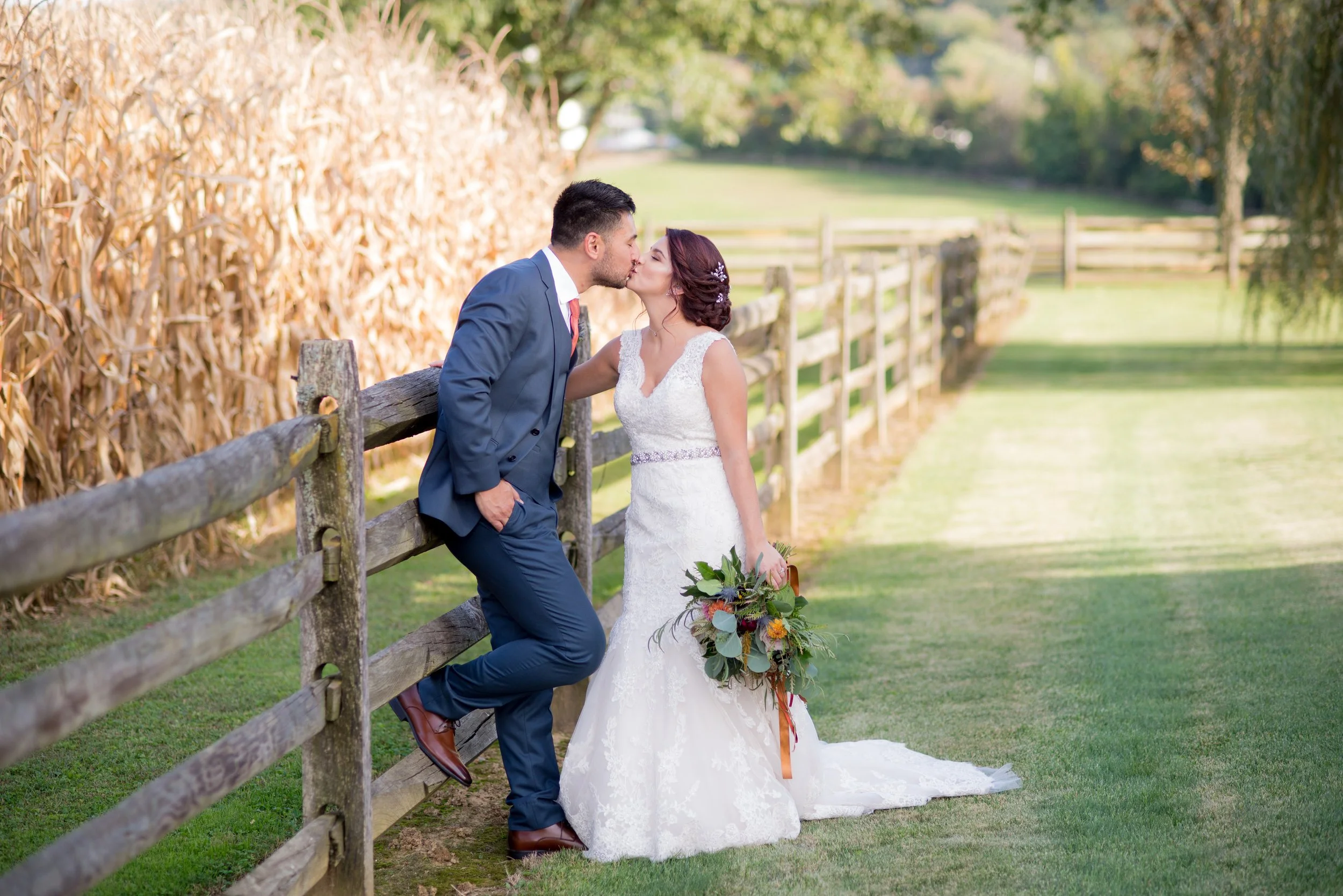 A bride and groom kissing outdoors in a rural setting, with the groom leaning against a wooden fence and the bride holding a bouquet, surrounded by green grass and trees.