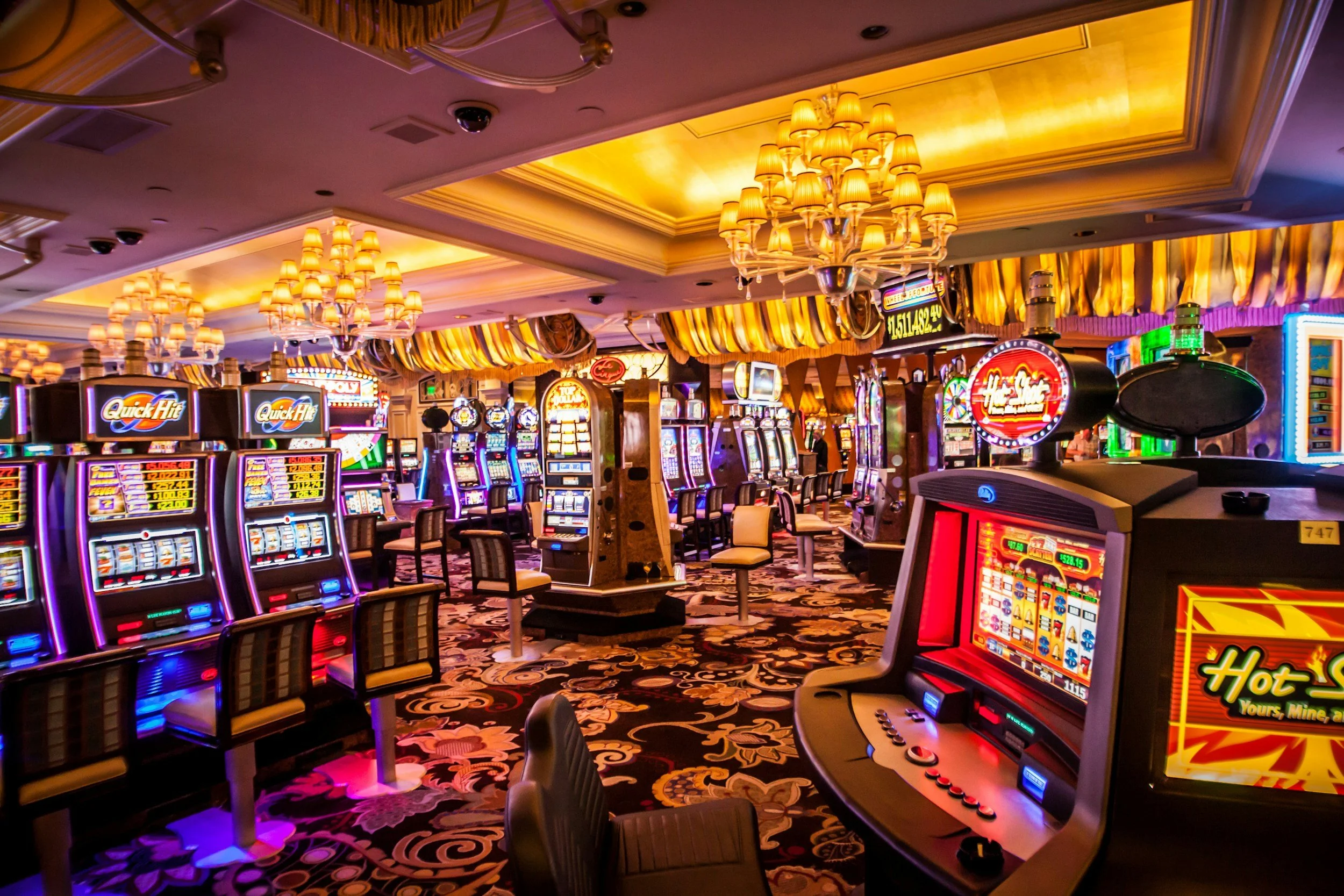 Interior of a casino filled with rows of slot machines, plush chairs, decorative chandeliers, and ornate carpeting.