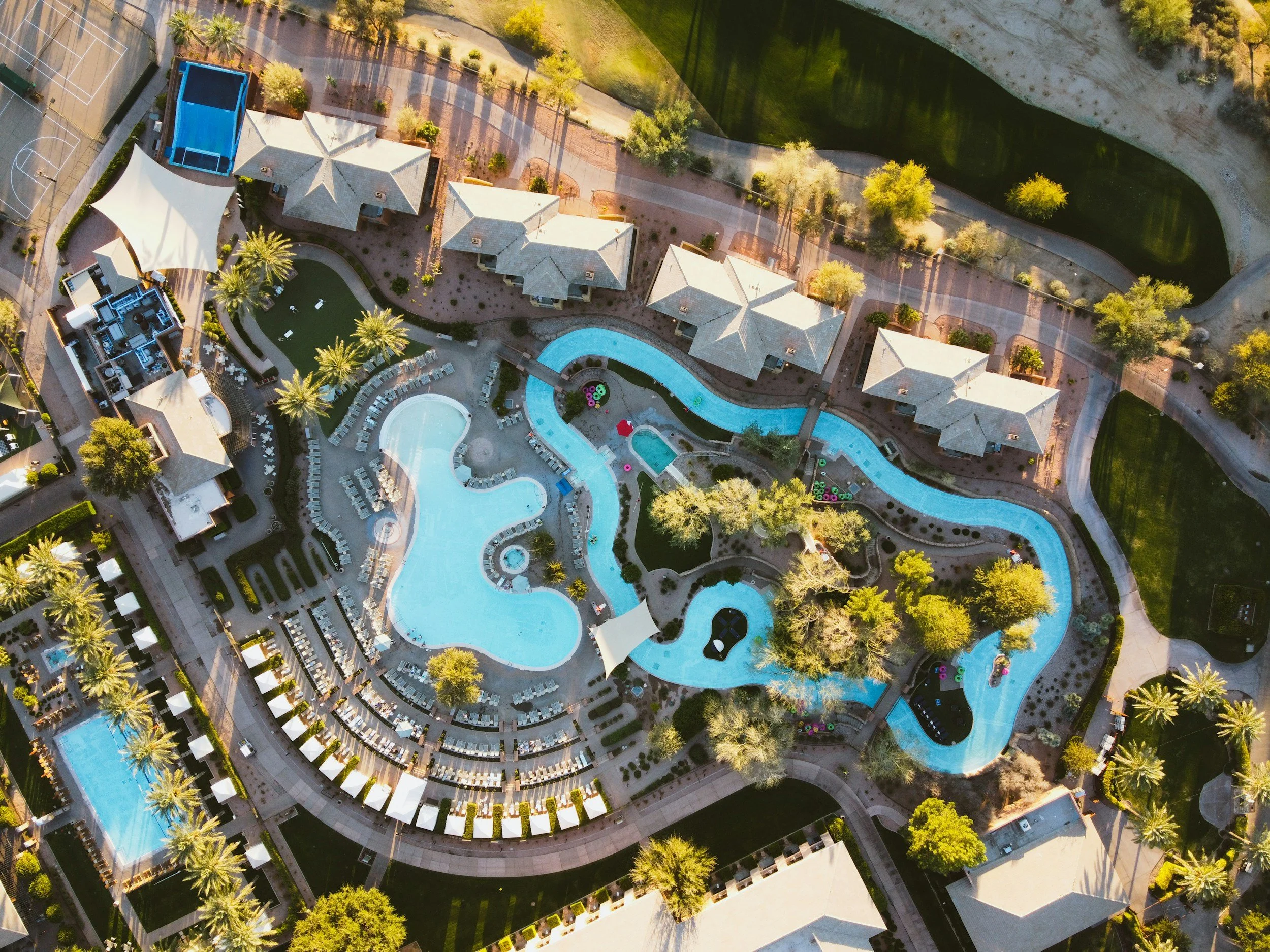 Aerial view of a large, multi-shape swimming pool complex at a resort, surrounded by lounge chairs, cabanas, palm trees, and adjacent to a grassy area and pathways.