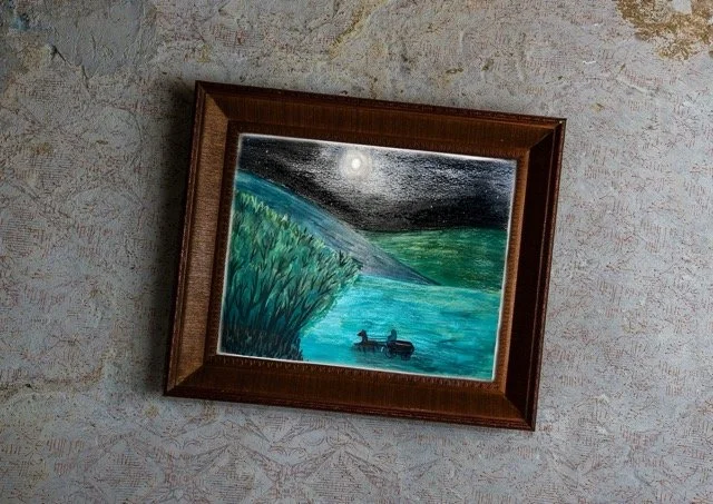 Framed landscape painting of a lake at night with a moon, grassy hills, and a boat on the water, hanging on a textured wall.