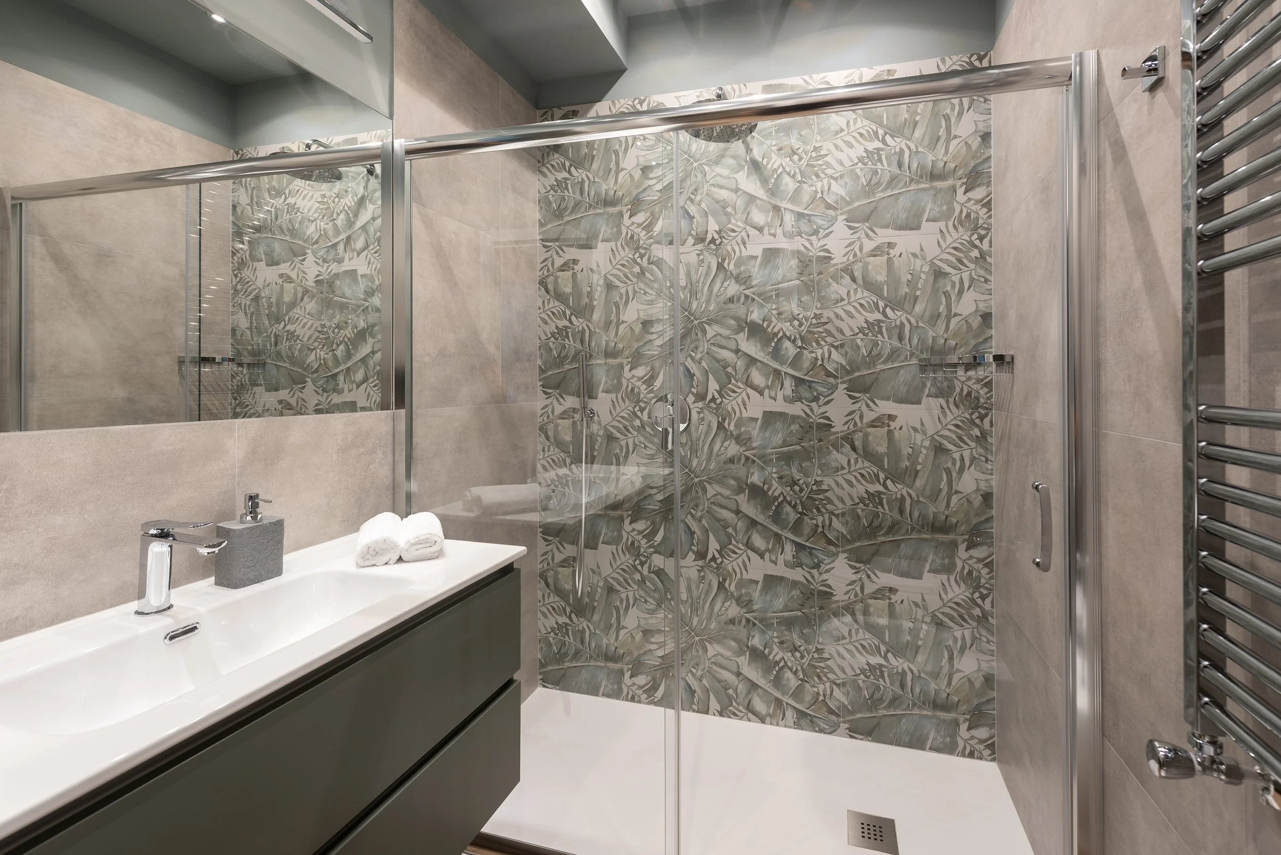 Modern bathroom with a glass-enclosed shower featuring tropical leaf patterned tiles, a sleek gray vanity with a white sink, two rolled-up white towels, and a chrome towel warmer.