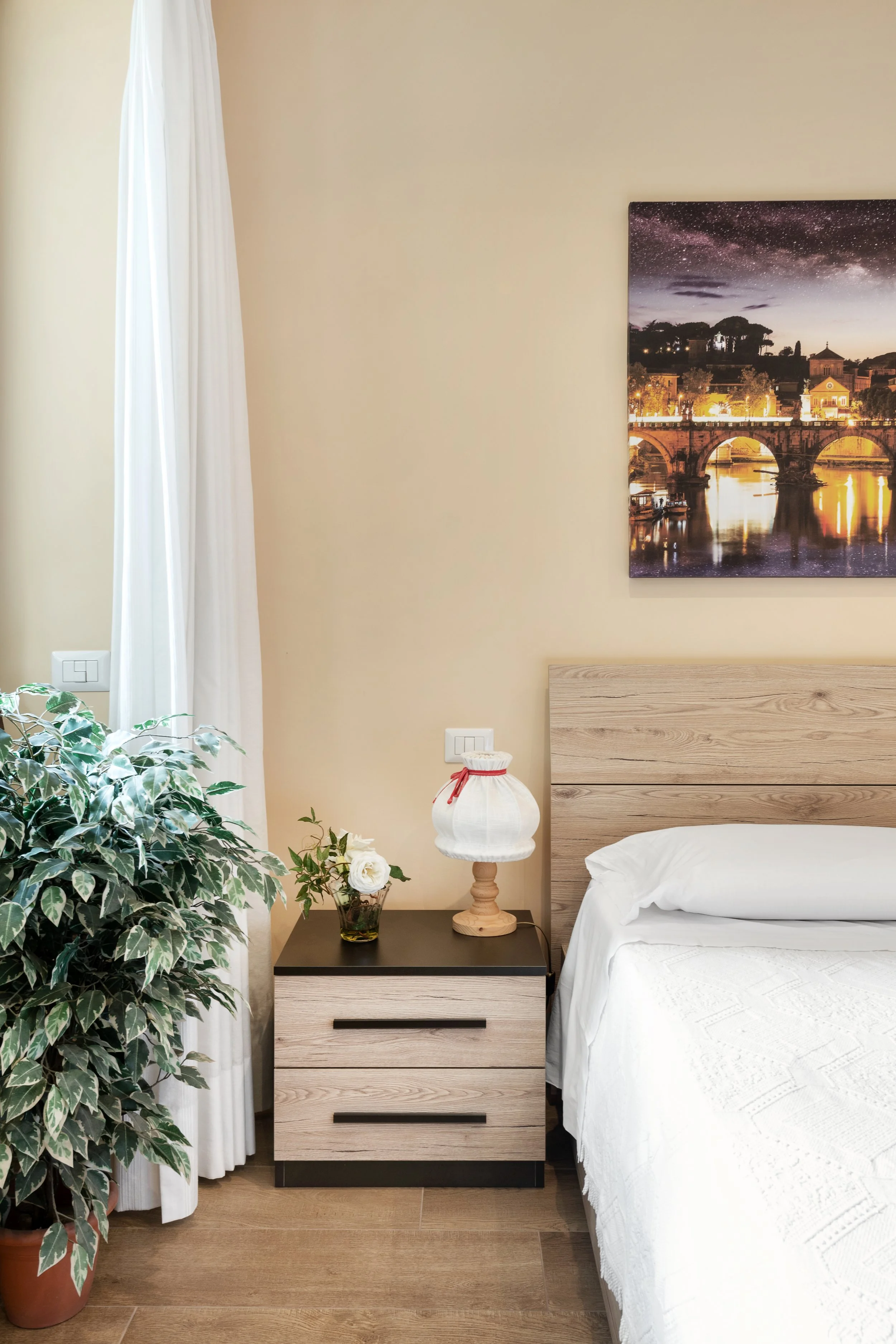 Bedroom with a beige wall, a nightstand with a flower vase and a table lamp, a large leafy plant, a window with white curtains, a bed with white bedding, and a framed cityscape photograph hanging above the bed.