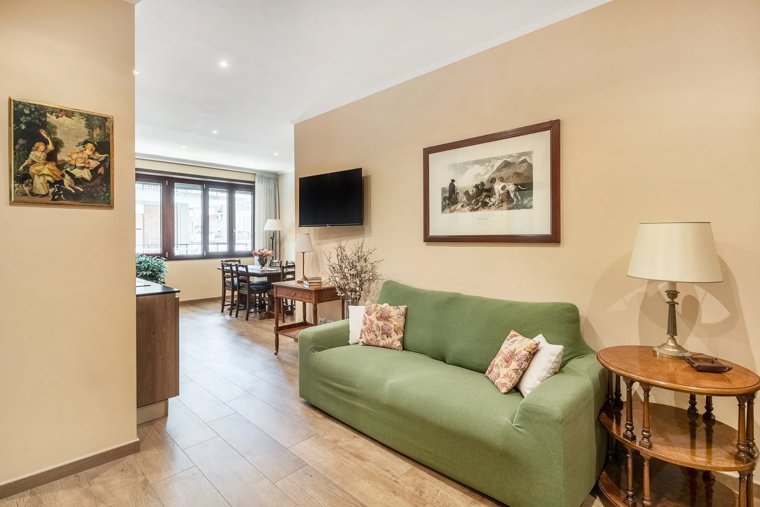 Living room featuring a green sofa with decorative pillows, a wooden side table with a lamp, wall art, a mounted flat-screen TV, and a dining area with a table and chairs near large windows.