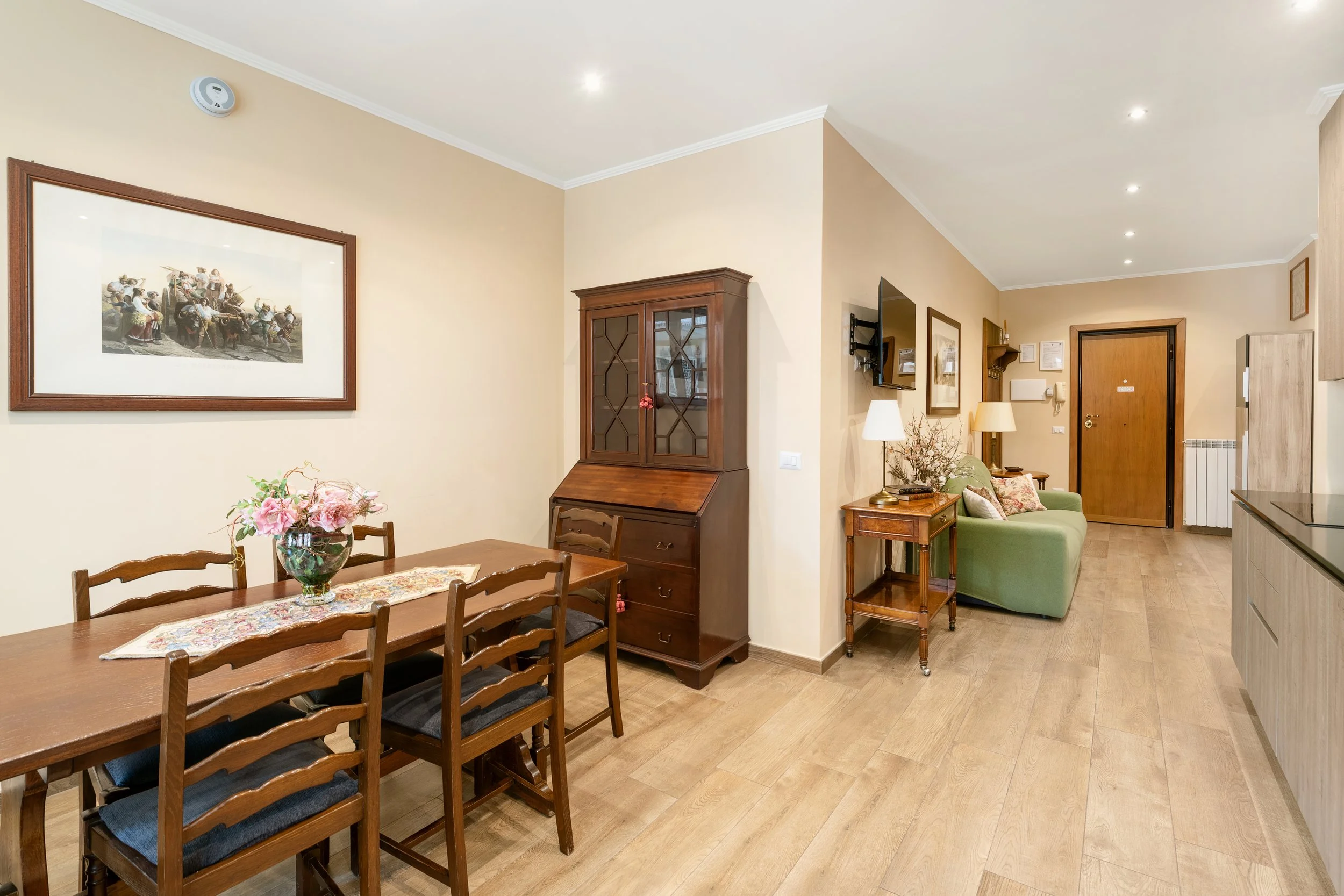 Living and dining area with hardwood flooring, wooden dining table with six chairs, glass vase with pink flowers, vintage wooden cabinet, green sofa with pillows, wall-mounted TV, framed artwork, table lamps, and a wooden front door.