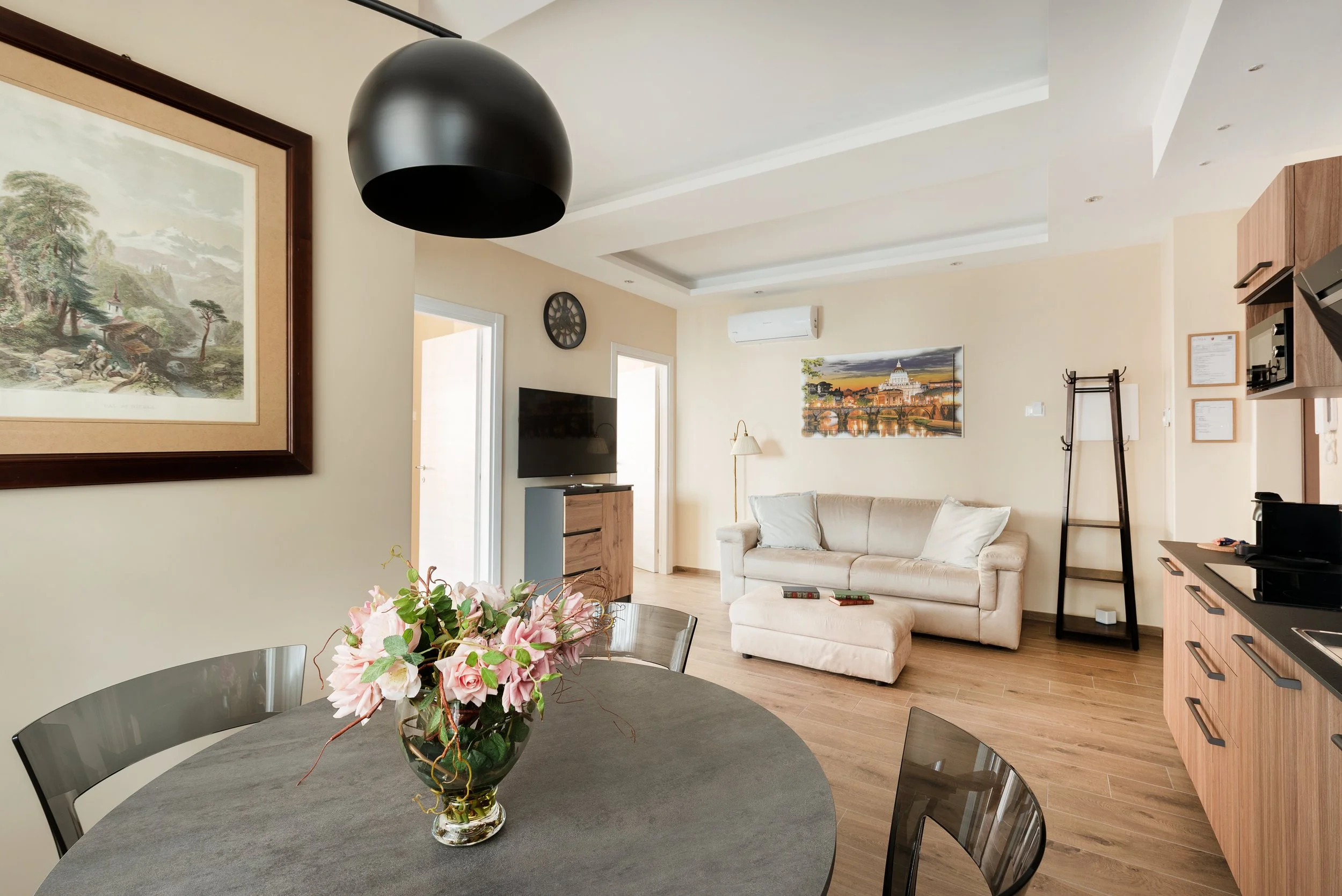 Living room with a dining table, floral centerpiece, beige sofa, wall art, TV, painting of a cityscape, wood flooring, and black ceiling light.