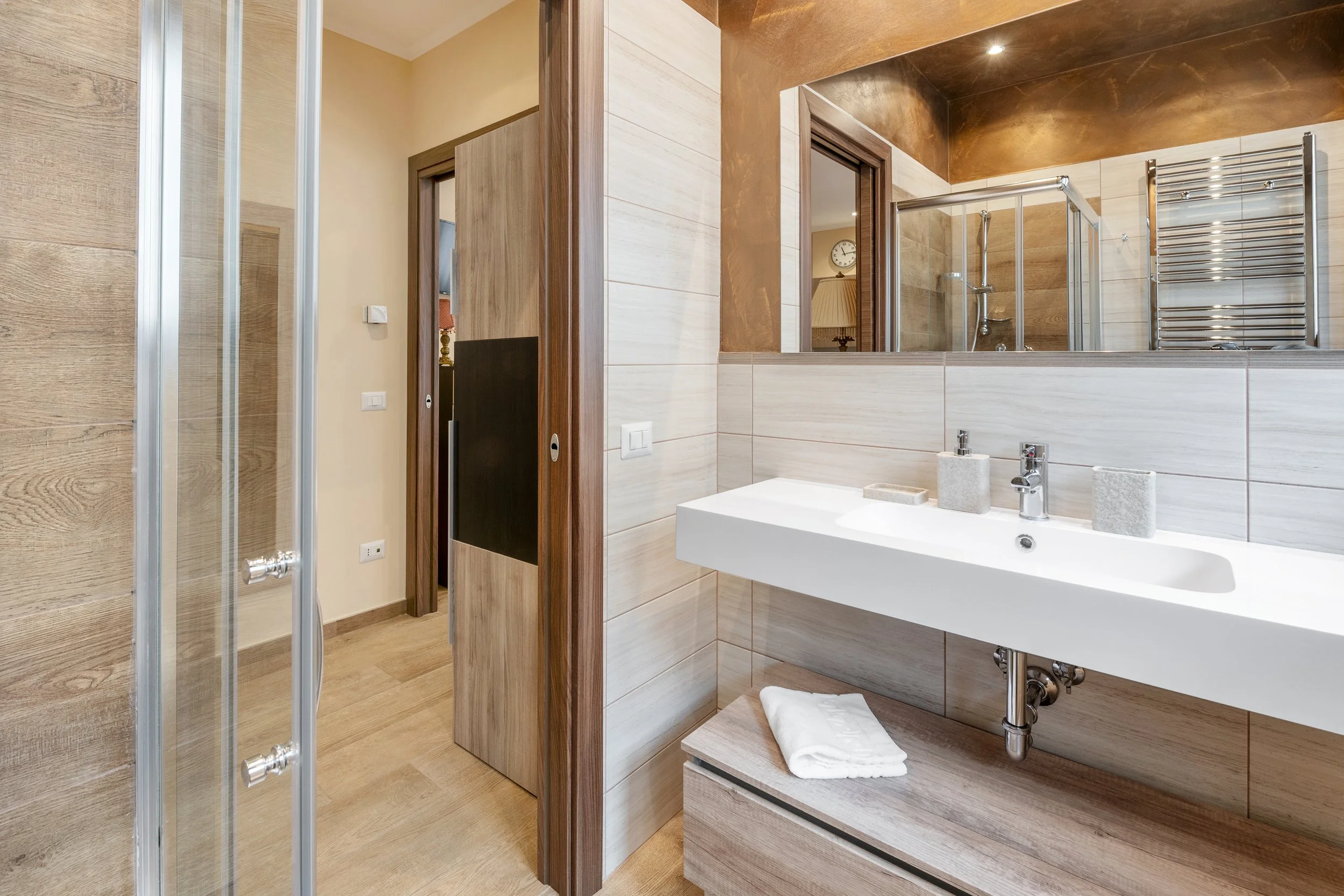 Modern bathroom with a white sink, wooden accents, a mirror, and a shower area with a glass door, towel rack, and wall clock visible through the mirror.