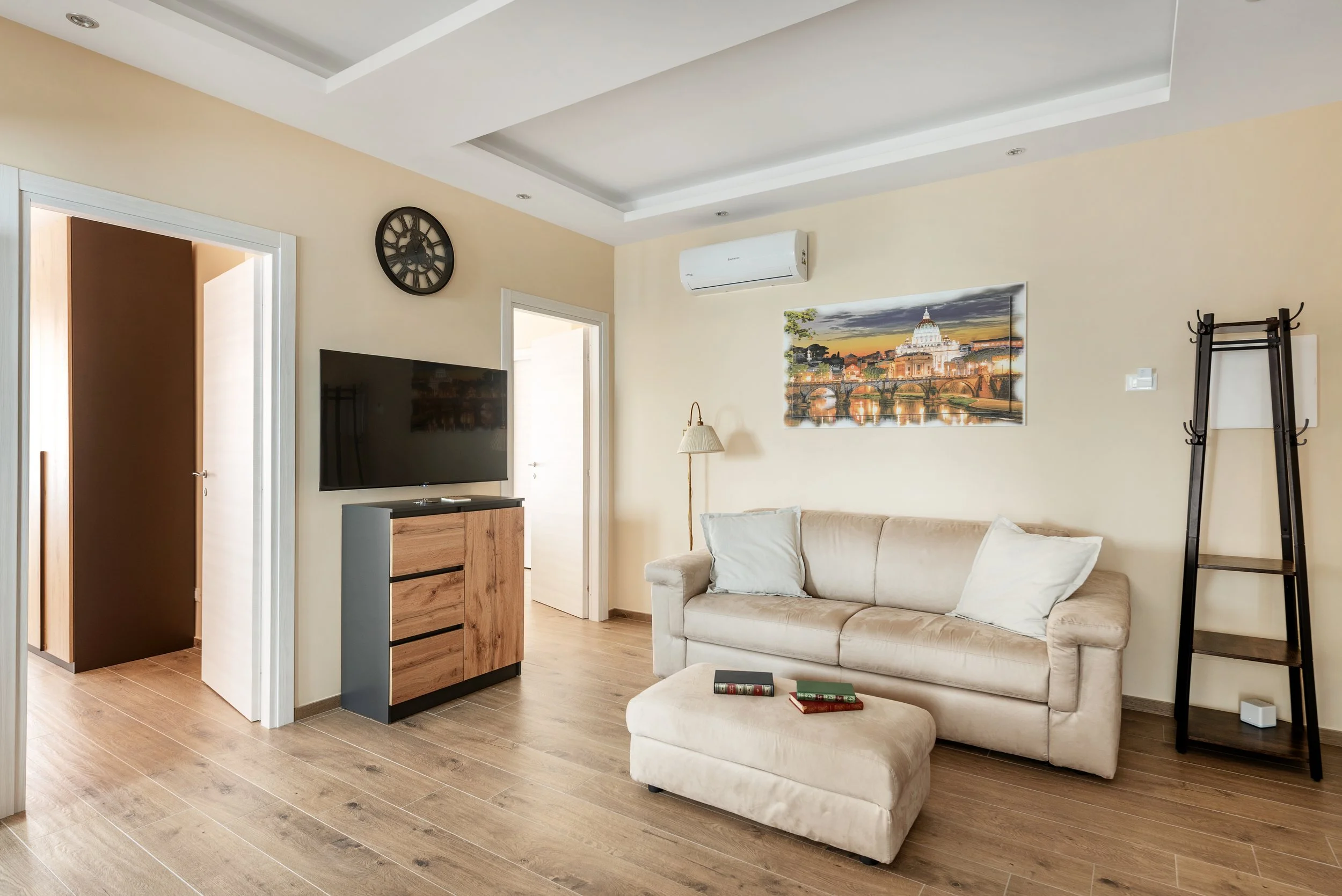 Living room with beige sofa, ottoman, wall art of European cityscape, wall clock, TV on wooden stand, wood floor, doorway, air conditioning unit, standing lamp, black ladder shelf.