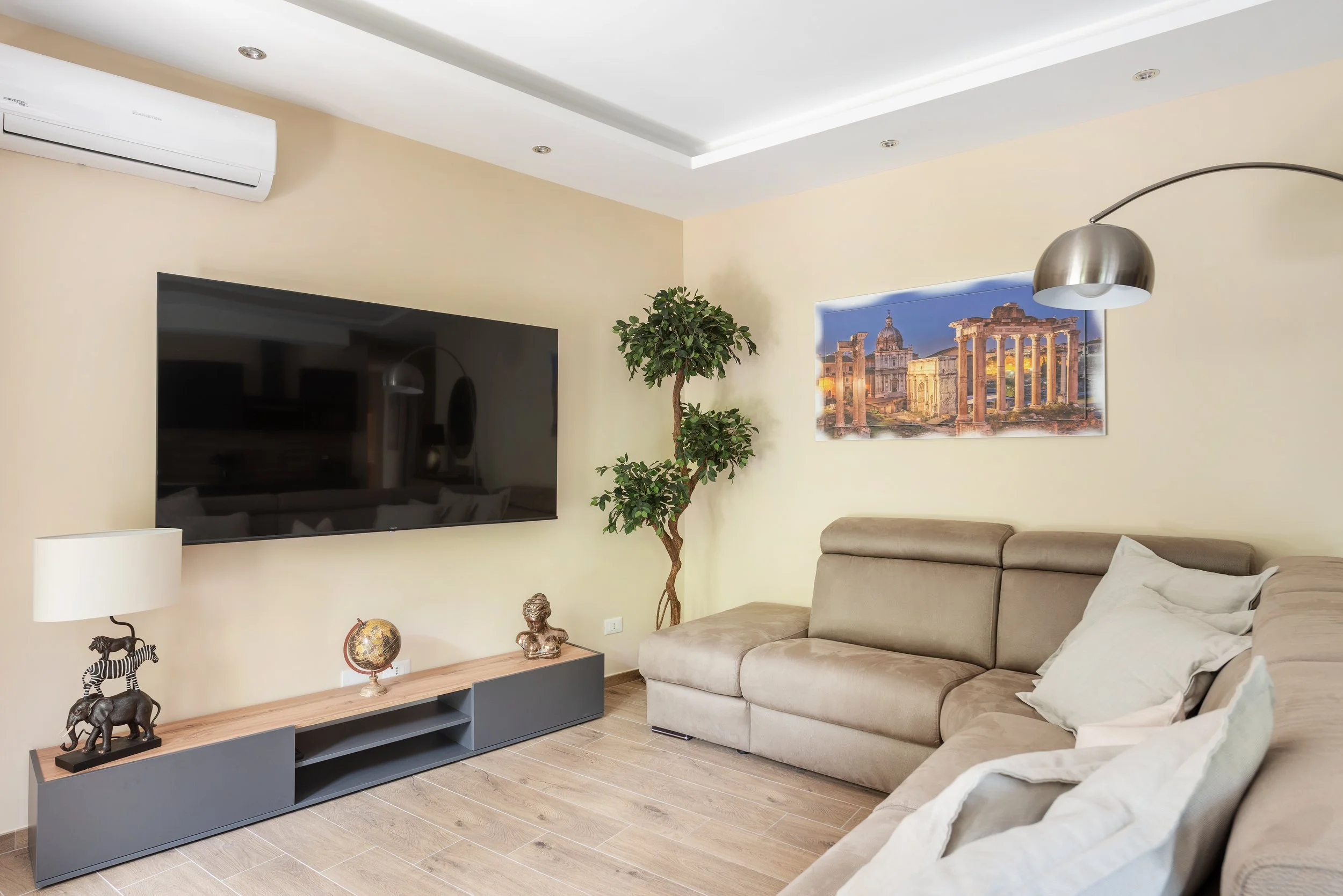 Living room with beige sectional sofa, wall-mounted flat-screen TV, potted tree, framed Roman forum scenery, and decorative items on a wooden TV stand.