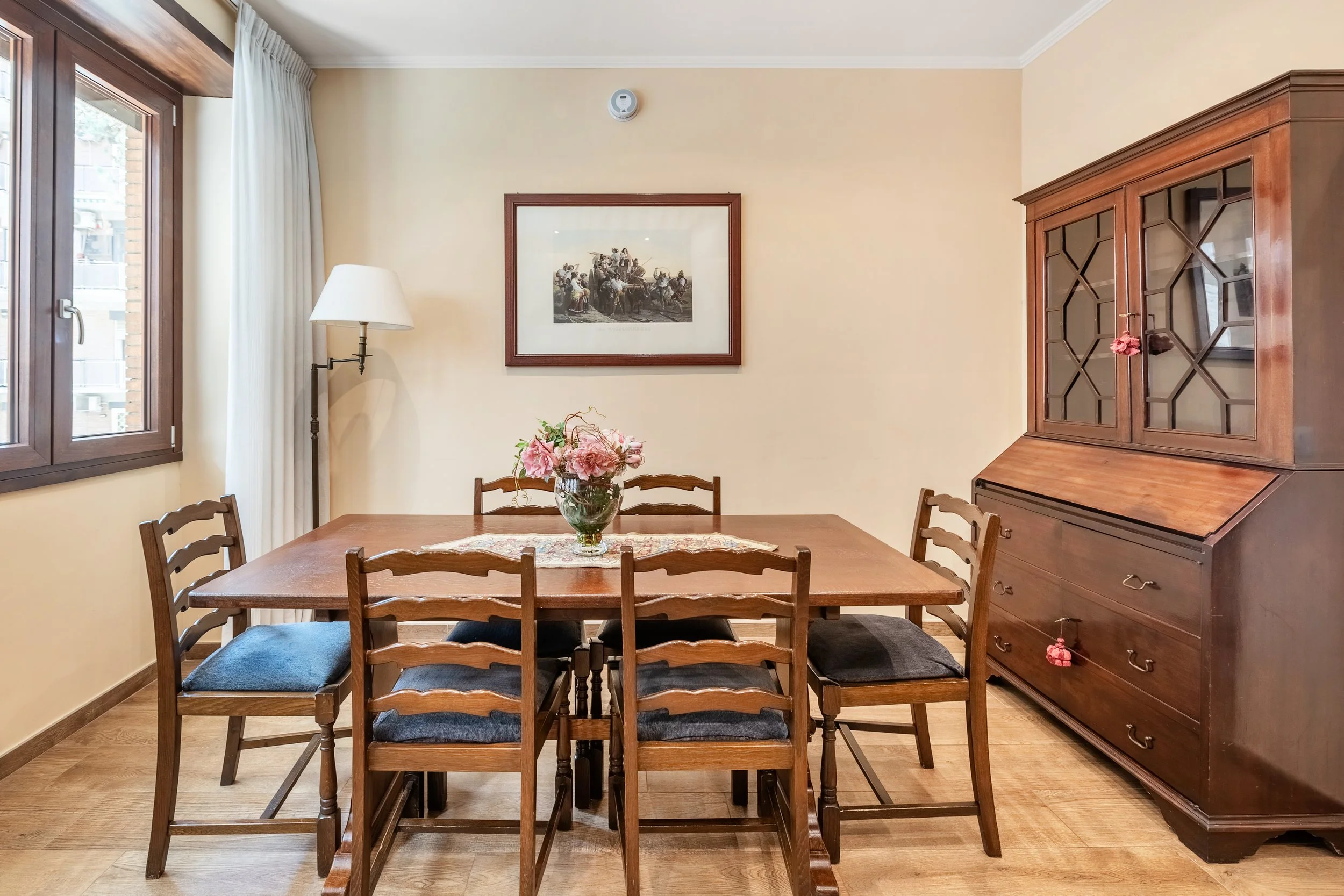 A dining room with a wooden table surrounded by six chairs, a vase with pink flowers on the table, a framed picture on the wall, a window with curtains, a floor lamp, and a wooden cabinet with glass doors and drawers.
