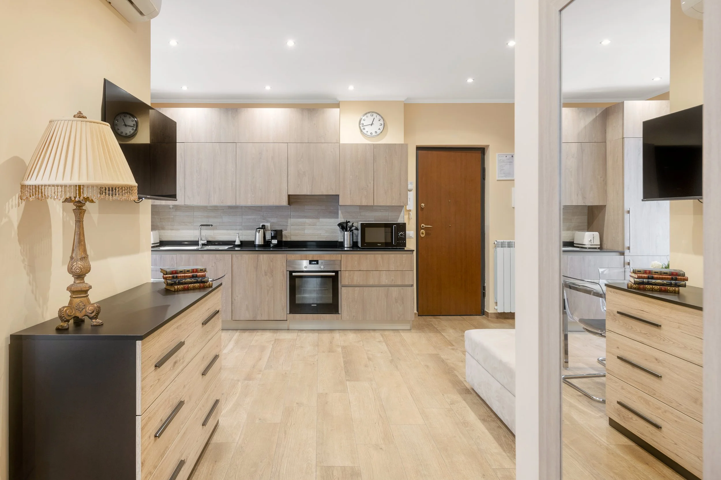 Modern kitchen with light wooden cabinets, black countertop, and stainless steel appliances. There is a wall clock, microwave, toaster, and kettle. A side table with a lamp and decorative items is visible, along with a mirror reflecting part of the r