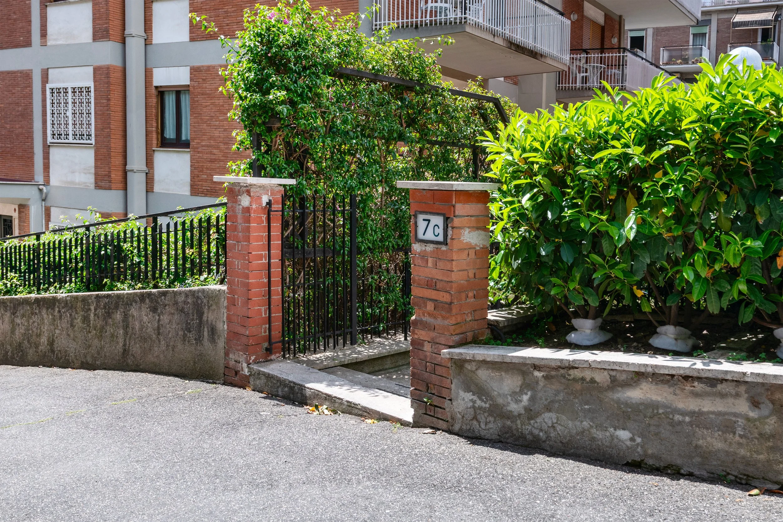 A brick and metal gate entrance to a residential building with a house number 7c on a brick pillar, surrounded by green bushes and trees in an urban neighborhood.