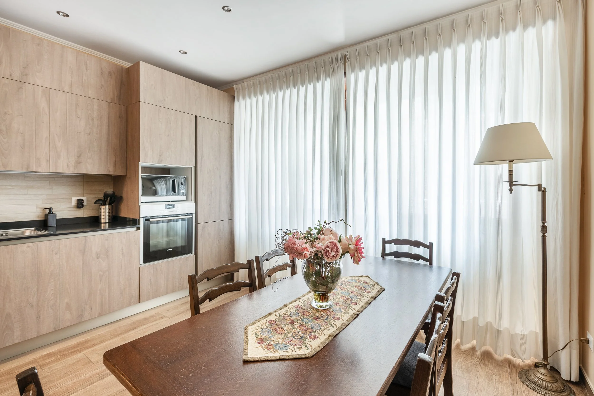 Dining room with wooden table, six chairs, a flower vase, and a window with white curtains, next to a built-in kitchen with wood cabinets, a microwave, and a floor lamp.