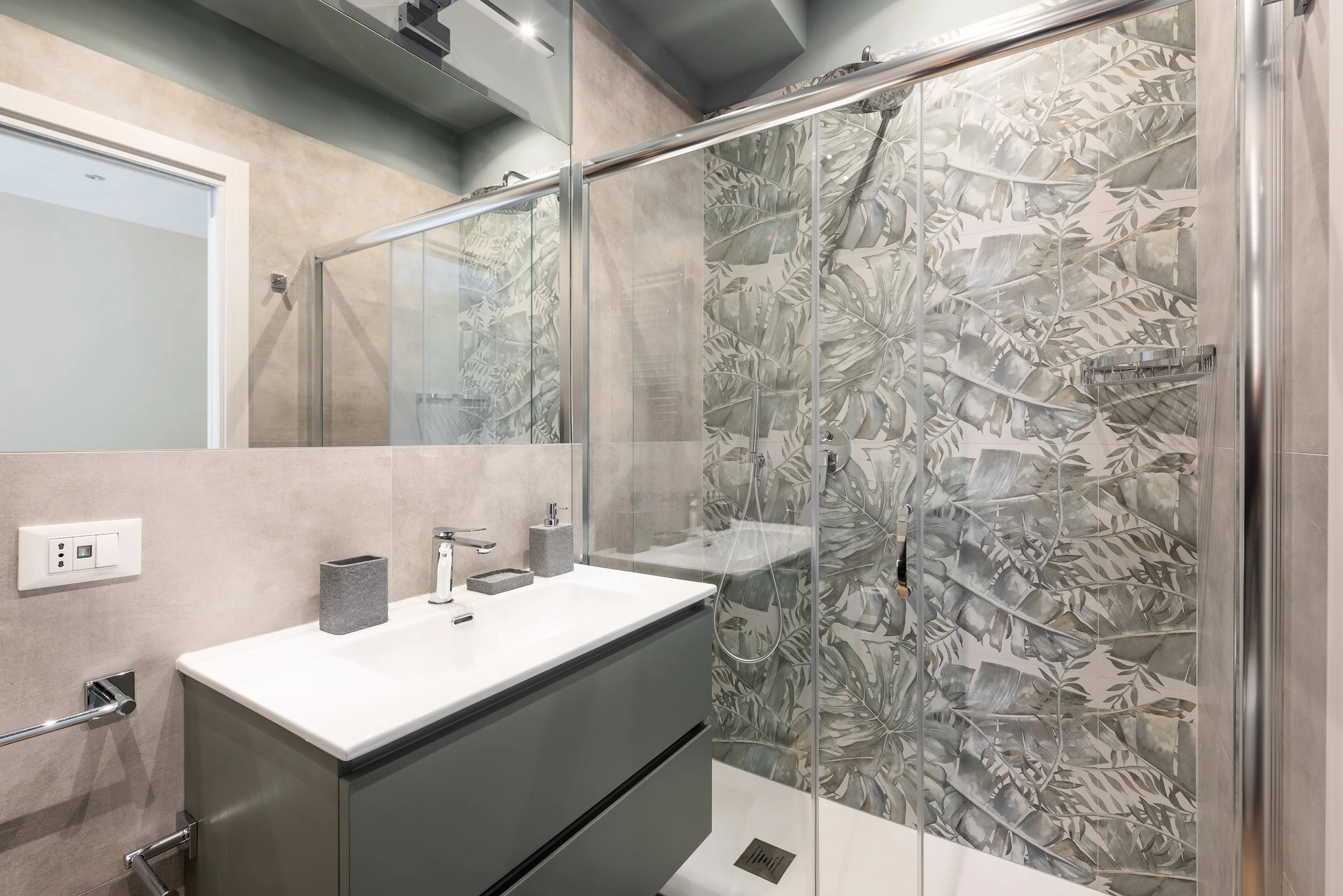 Modern bathroom with a gray vanity, a large mirror, a textured wallpaper with large tropical leaves, and a glass-enclosed shower.