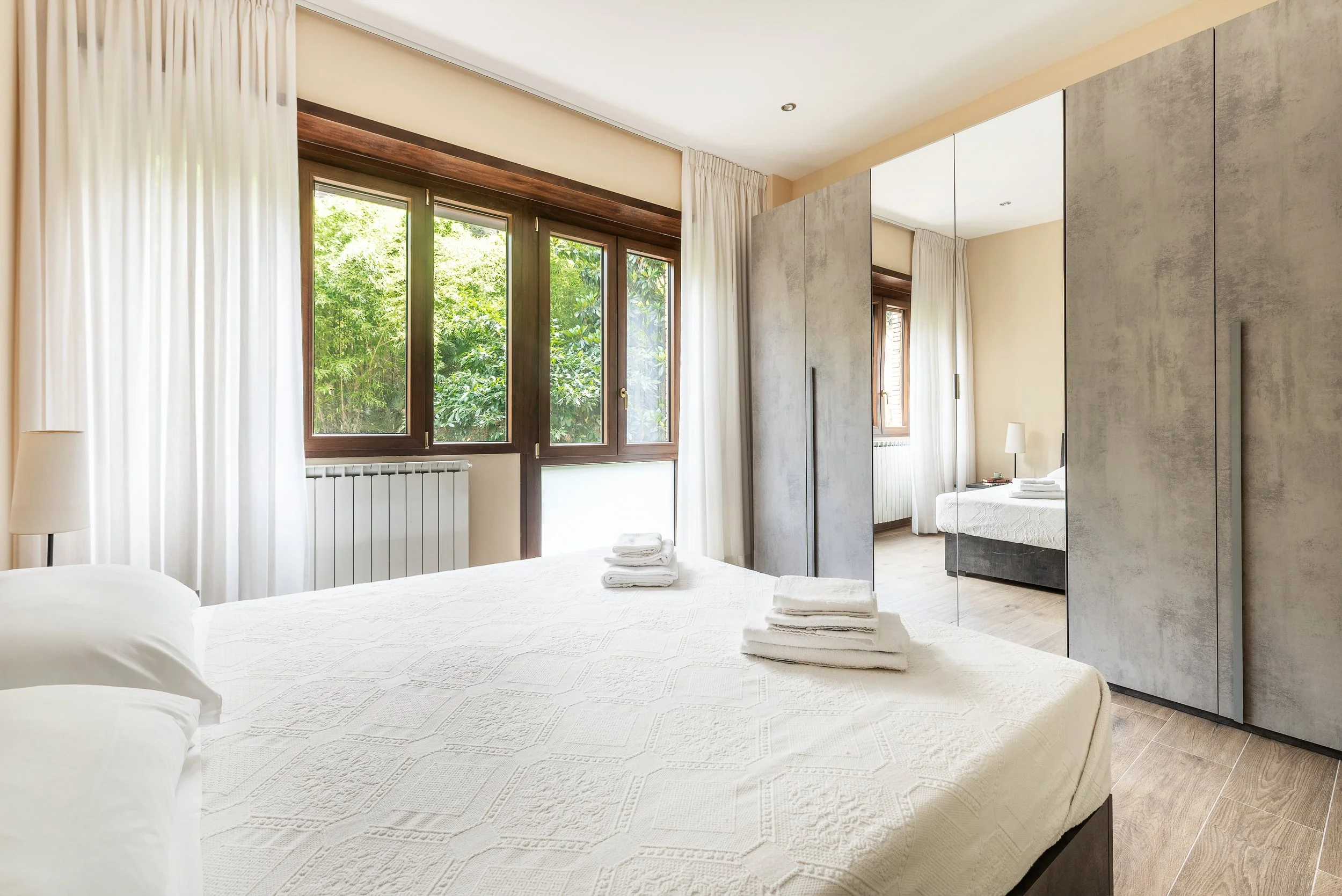 Bright bedroom with large windows, white bed, folded towels, and a wardrobe with mirrored doors.