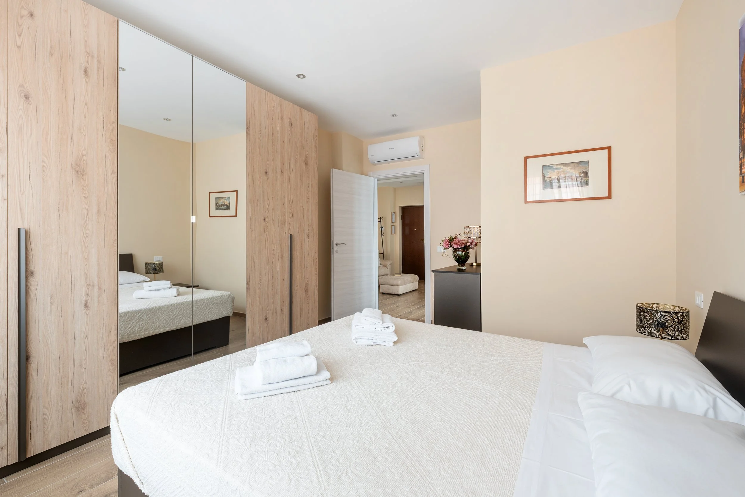 A tidy hotel bedroom with a large bed, mirrored wardrobe, framed pictures on the cream-colored walls, and an open door leading to a hallway with a chair and coat rack.