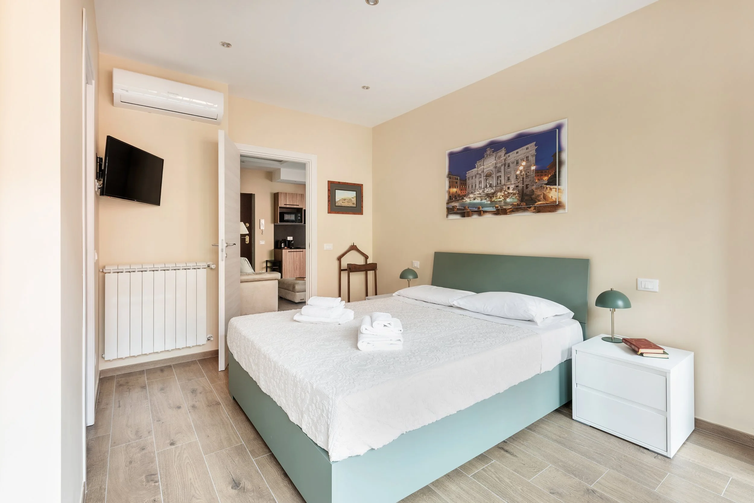 A bedroom with a bed, two bedside lamps, a painting above the bed, and a flat-screen TV on the wall. An open door reveals a sitting area with a sofa and kitchen in the background.