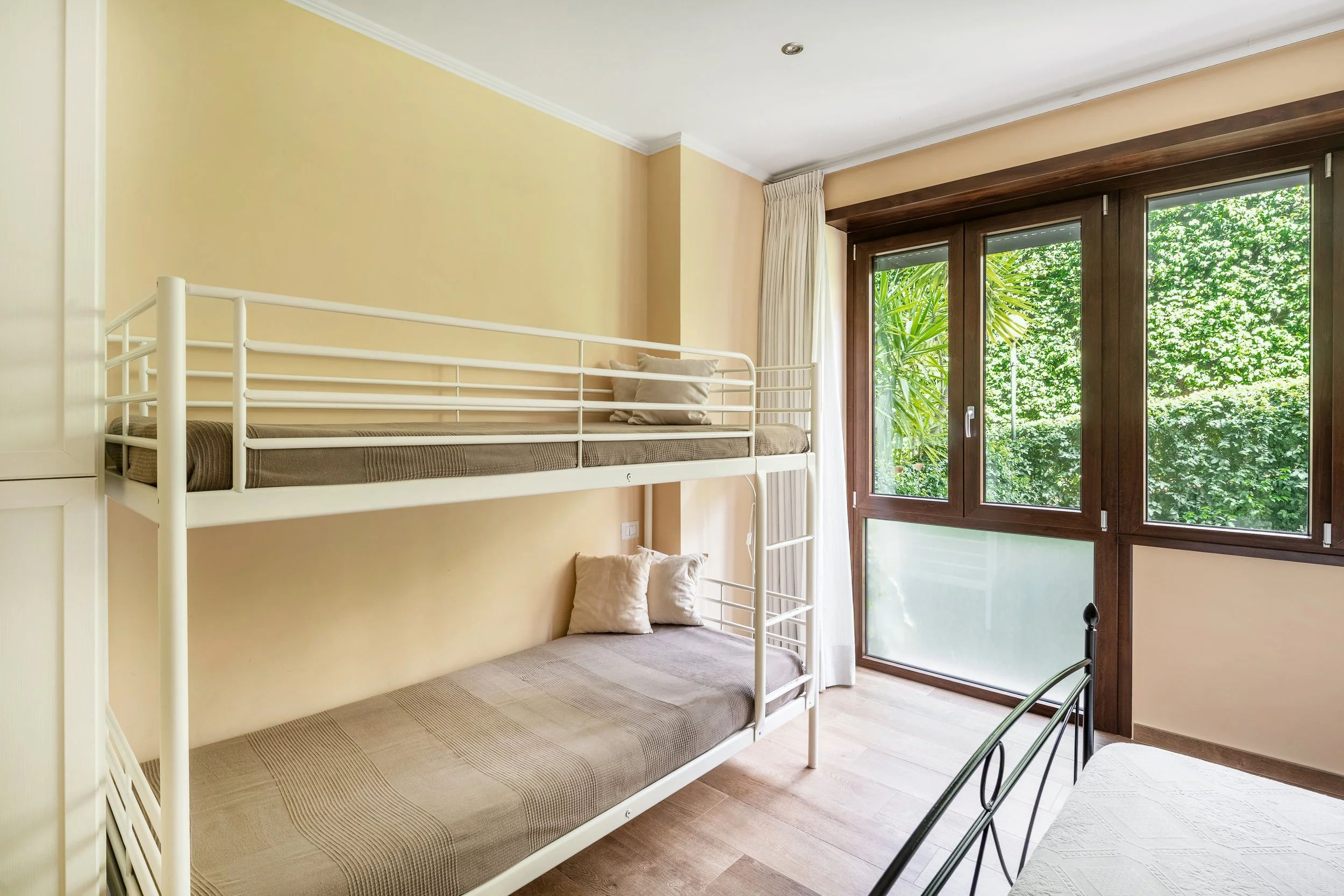 Room with bunk beds, window showing green trees, beige walls, and hardwood floors.
