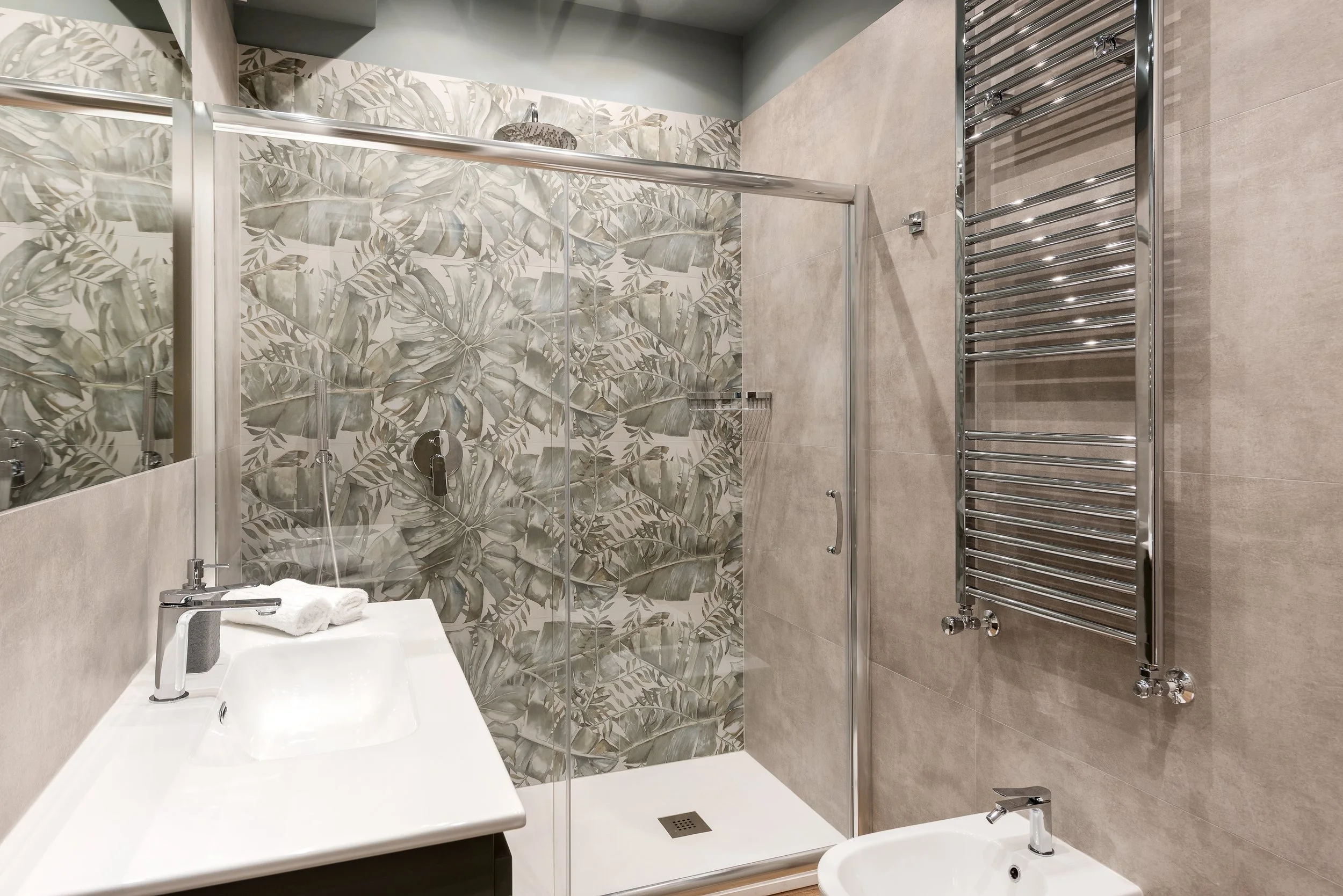 Modern bathroom with a walk-in shower that has a tropical leaf patterned tile wall, a white sink, a towel on the sink, a chrome towel warmer, and a bidet.