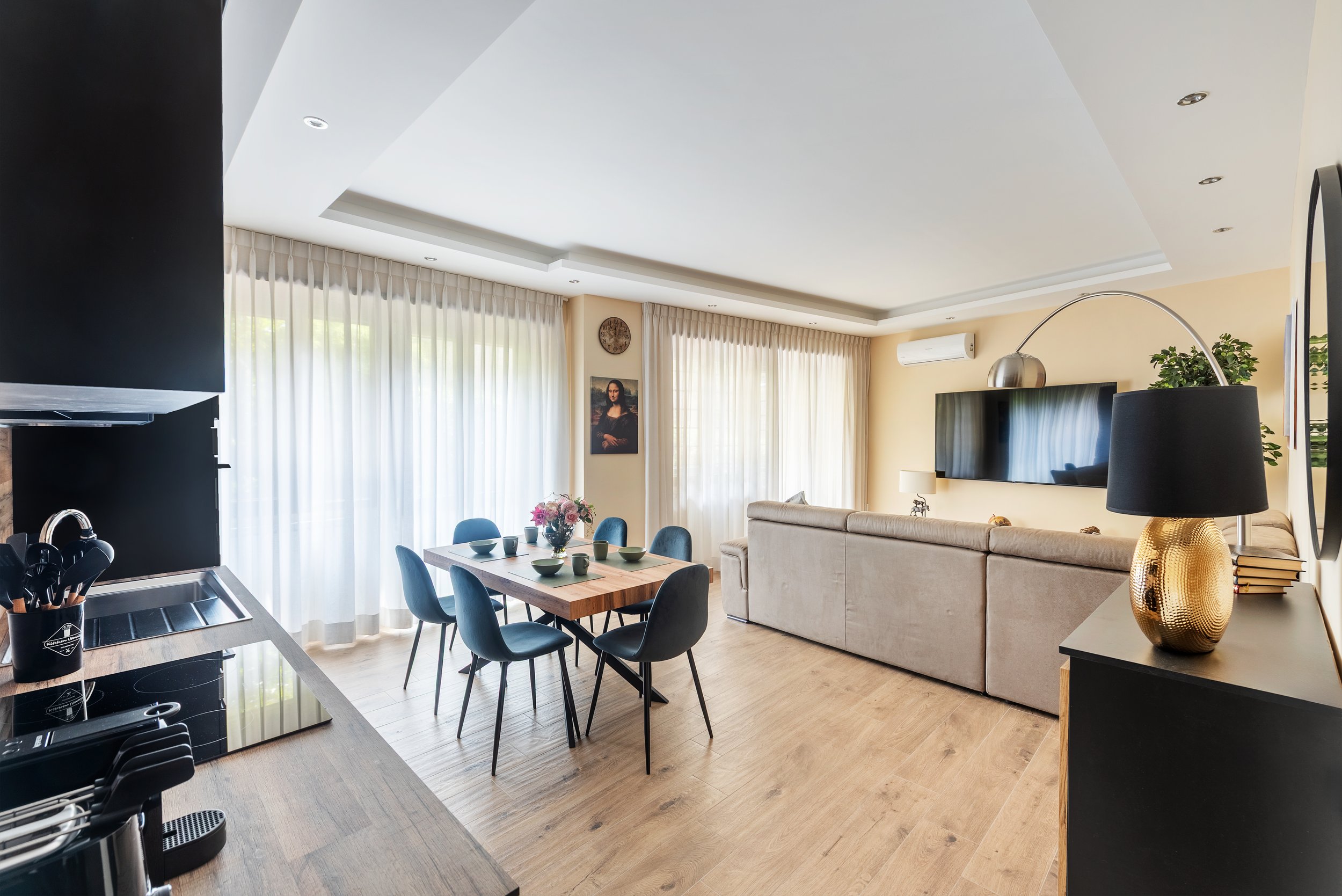 Modern living room with beige couch, wall-mounted TV, black side table with gold lamp, wooden dining table with blue chairs, large windows with sheer curtains, and wooden flooring.