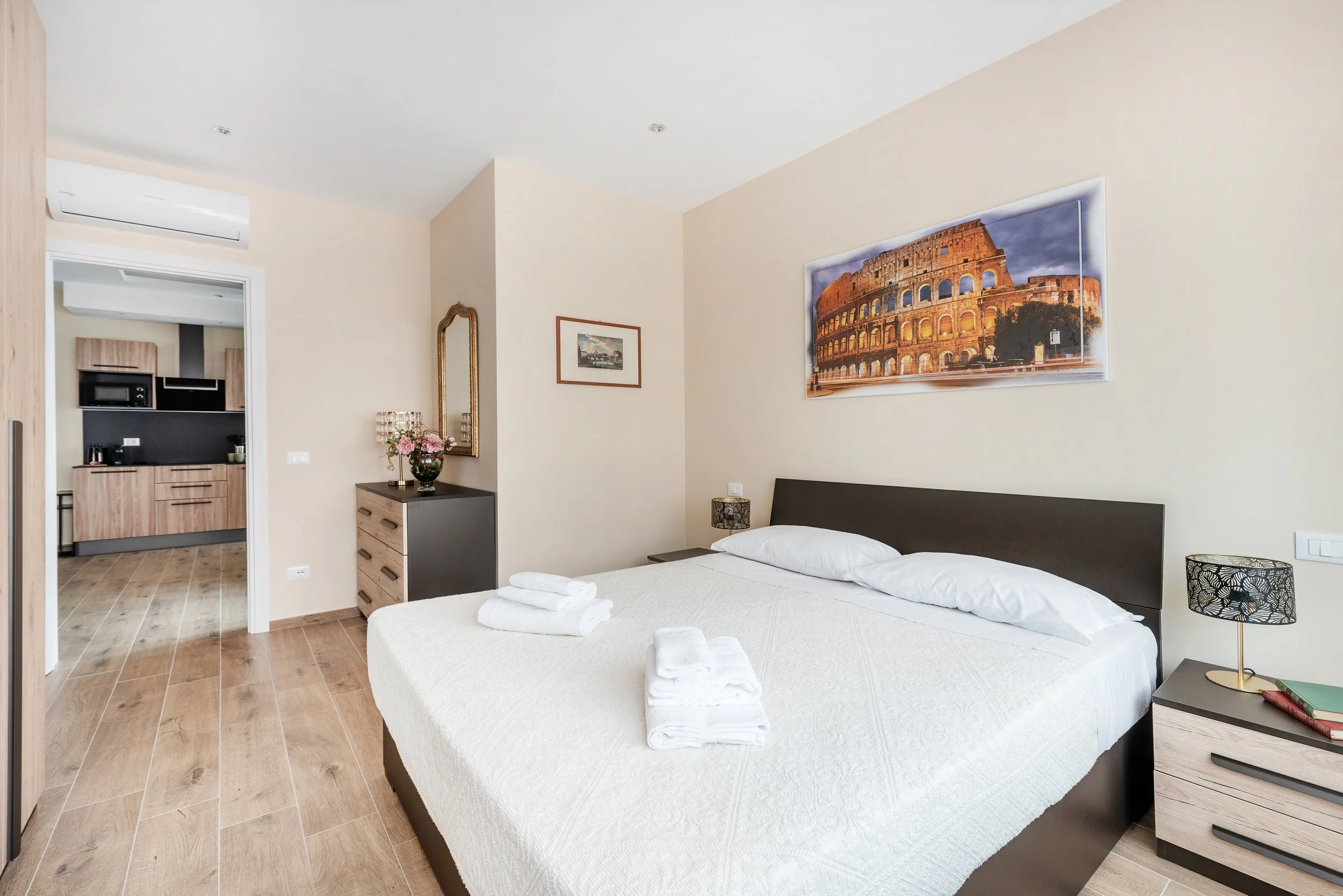 Bedroom with a large bed, nightstands, and wall art of the Colosseum. The room has light beige walls and wooden flooring, with a view into a kitchen area.