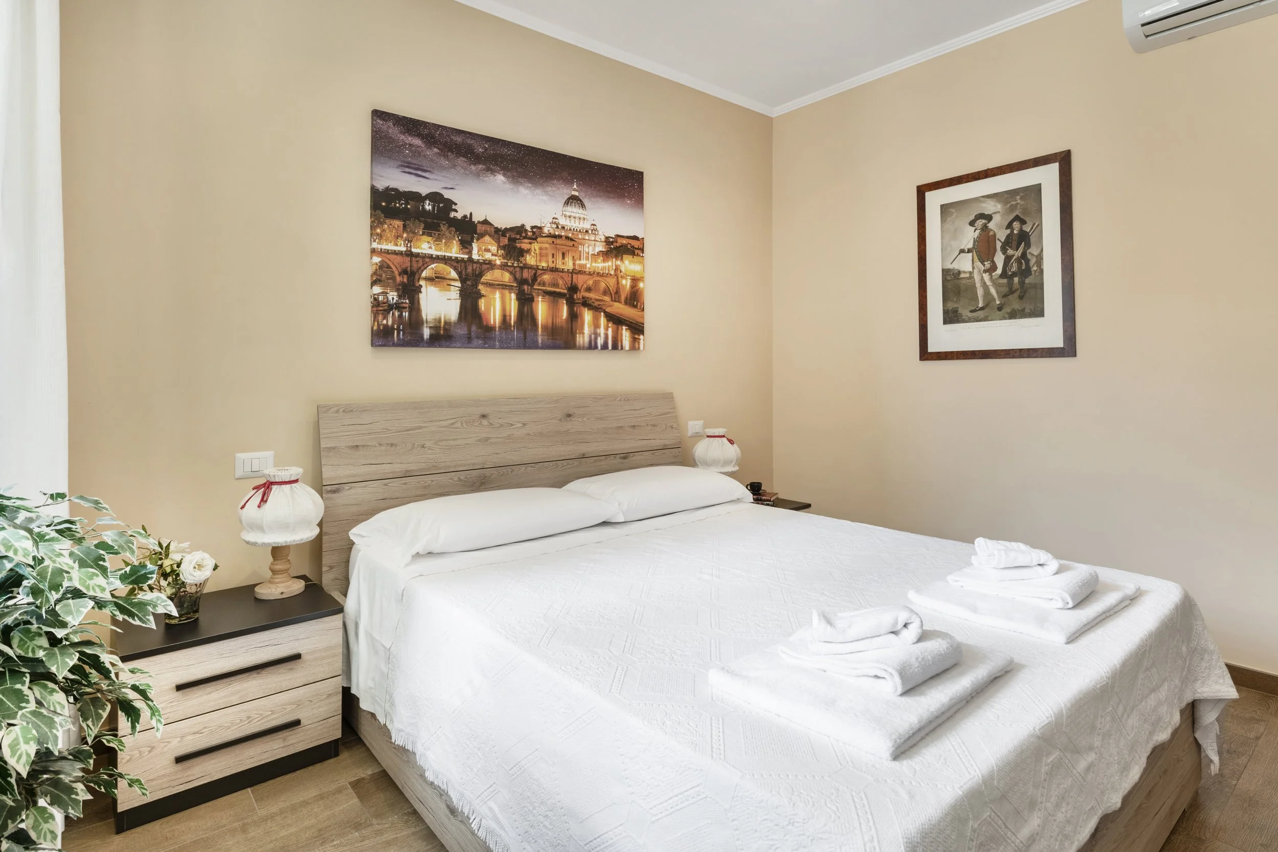 A neatly made bed with white linens and towels folded on top, in a room with beige walls and two framed pictures, one of Rome's cityscape and another of historical figures, with a wooden nightstand and a decorative plant beside the bed.