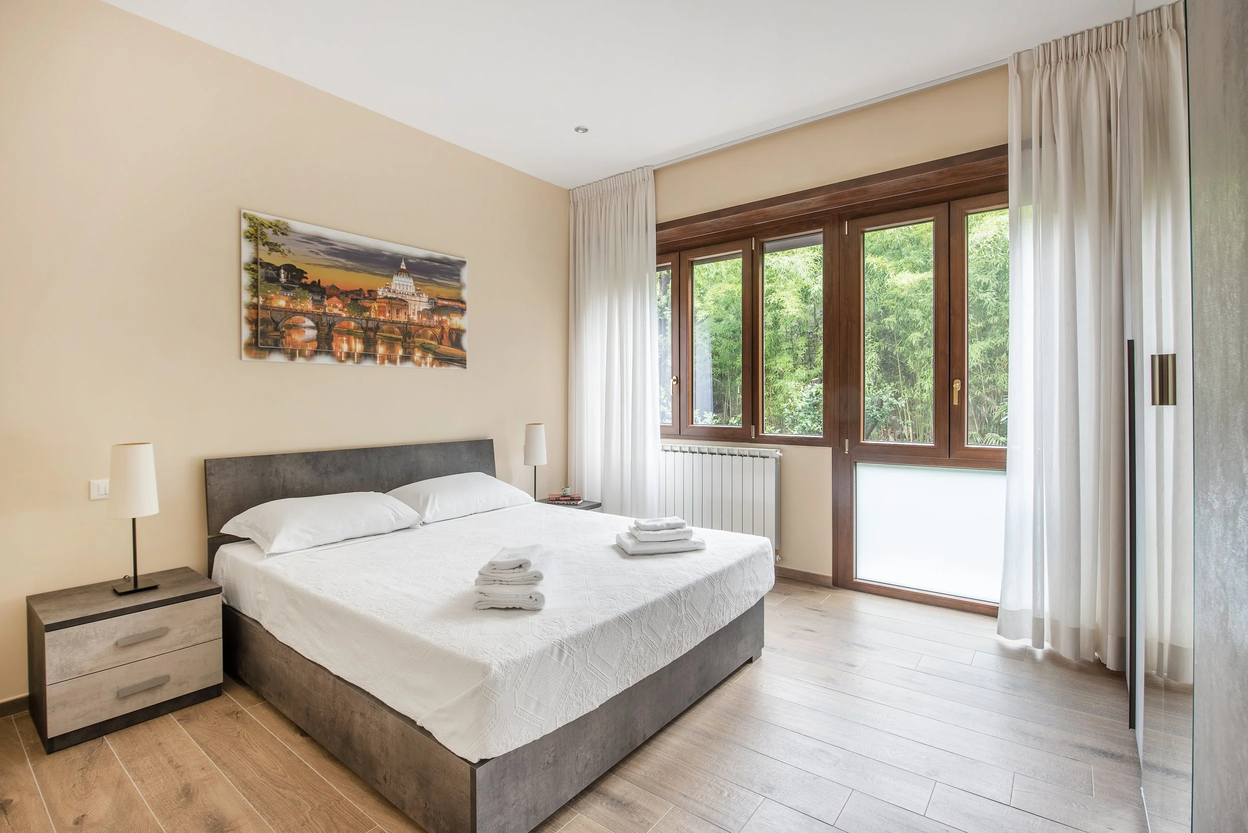 Bright hotel room with a large bed, white bedding, and two stacks of towels. Nightstands with lamps on each side of the bed. Large windows with white curtains showing green trees outside.