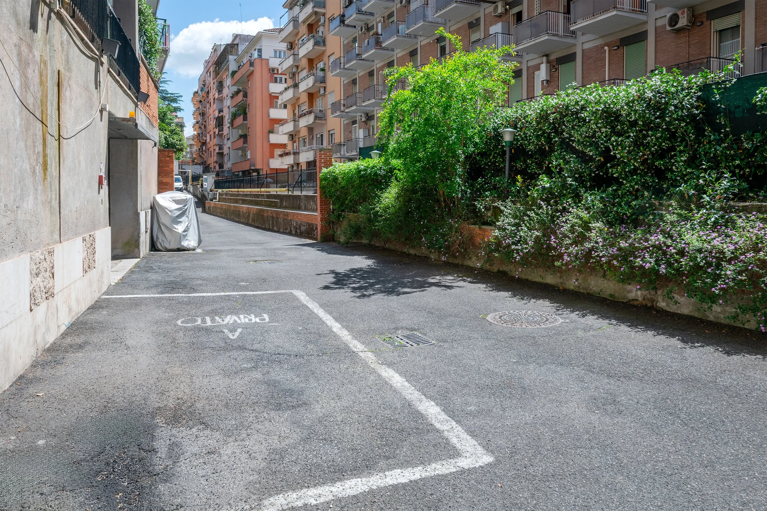 An empty parking space marked 'reserved' in an alley, with a covered bicycle or scooter next to a building wall on the left, and residential buildings with balconies and greenery on the right.