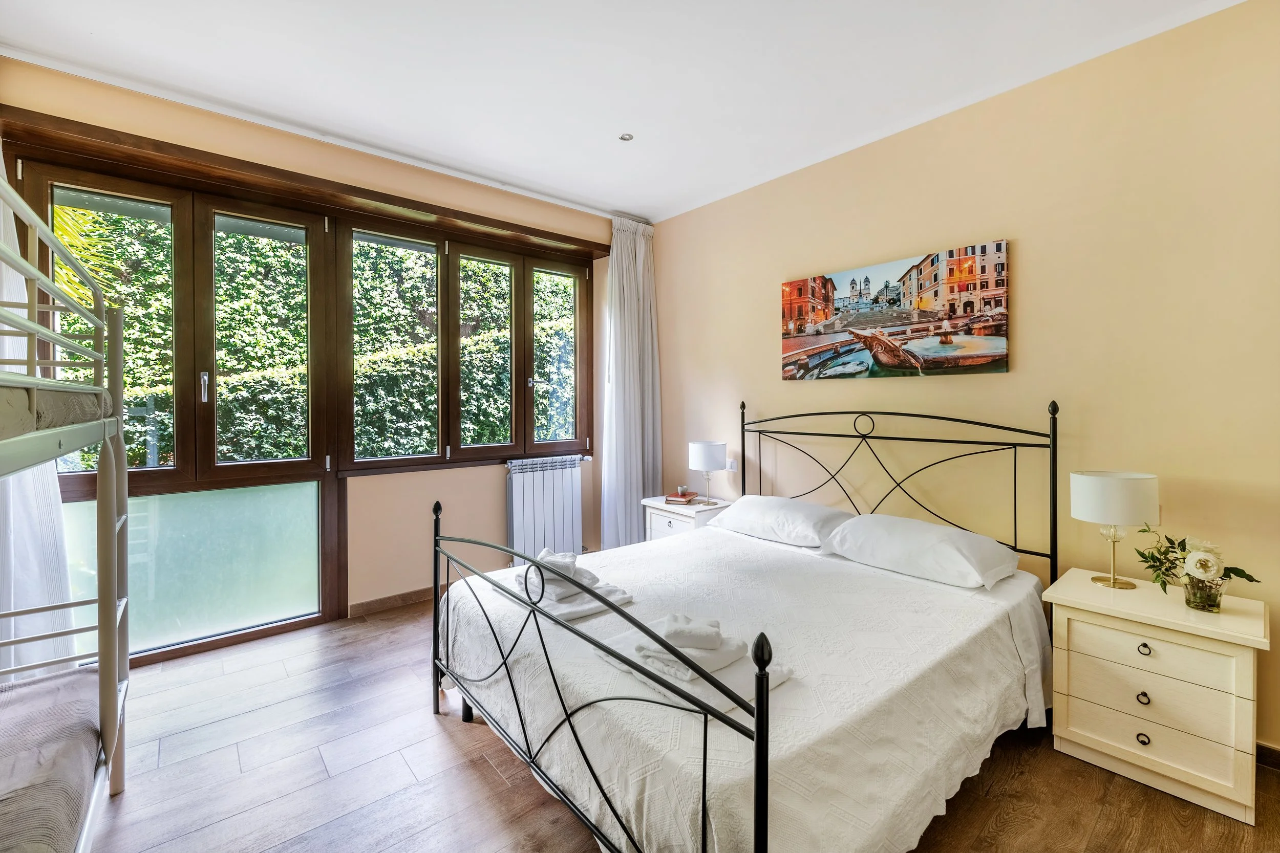 Hotel bedroom with large bed, nightstands, lamps, a floral arrangement, and large windows with green foliage outside.