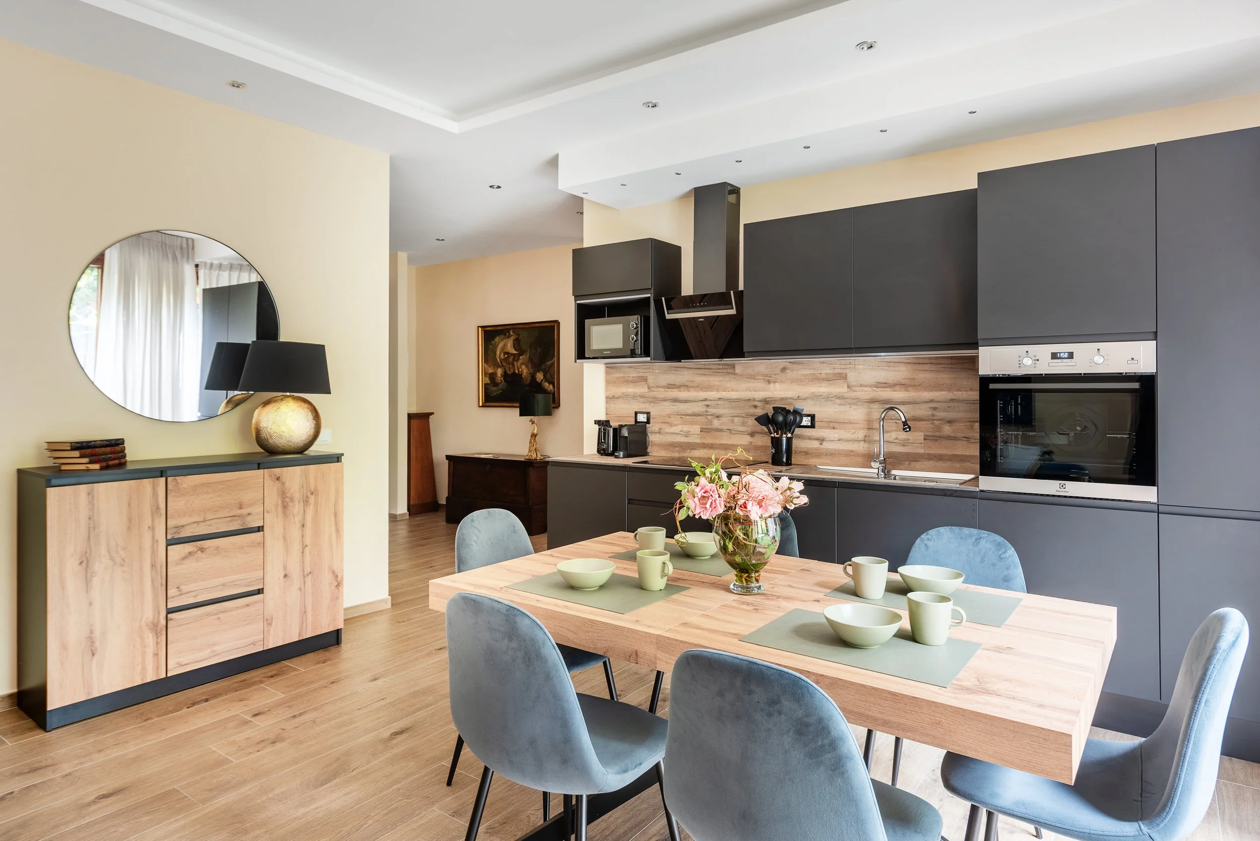 Modern kitchen and dining area with wooden dining table, six gray chairs, a floral centerpiece, black kitchen cabinets, built-in microwave and oven, wooden backsplash, and decorative items on a sideboard.