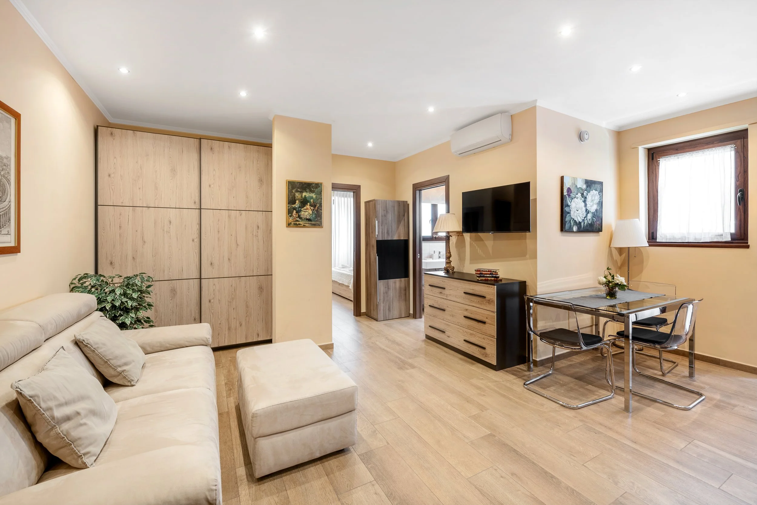 Living room with beige sofa, ottoman, wooden floor, wall art, and dining area with glass table and black chairs, window with curtains, and flat-screen TV.