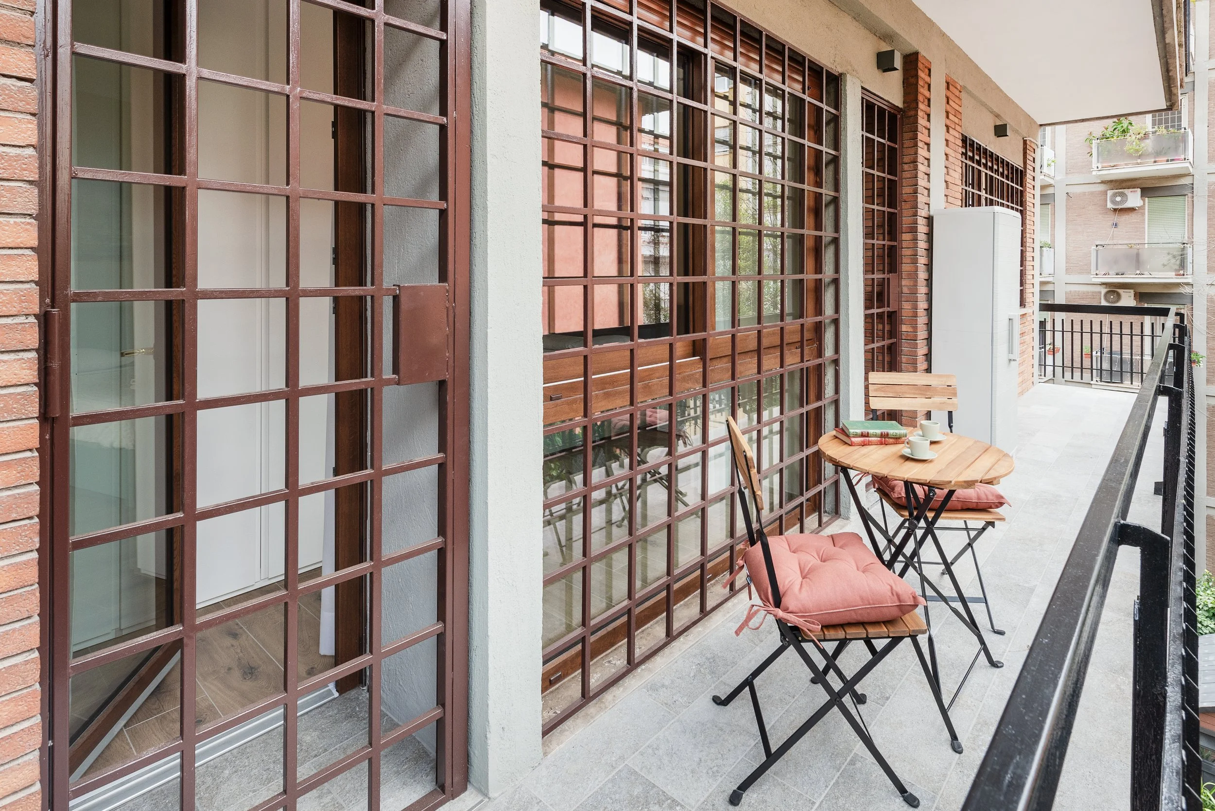 Balcony with a small round wooden table, two chairs with cushions, and a book, with large glass windows and metal grid gates, in an urban apartment building.