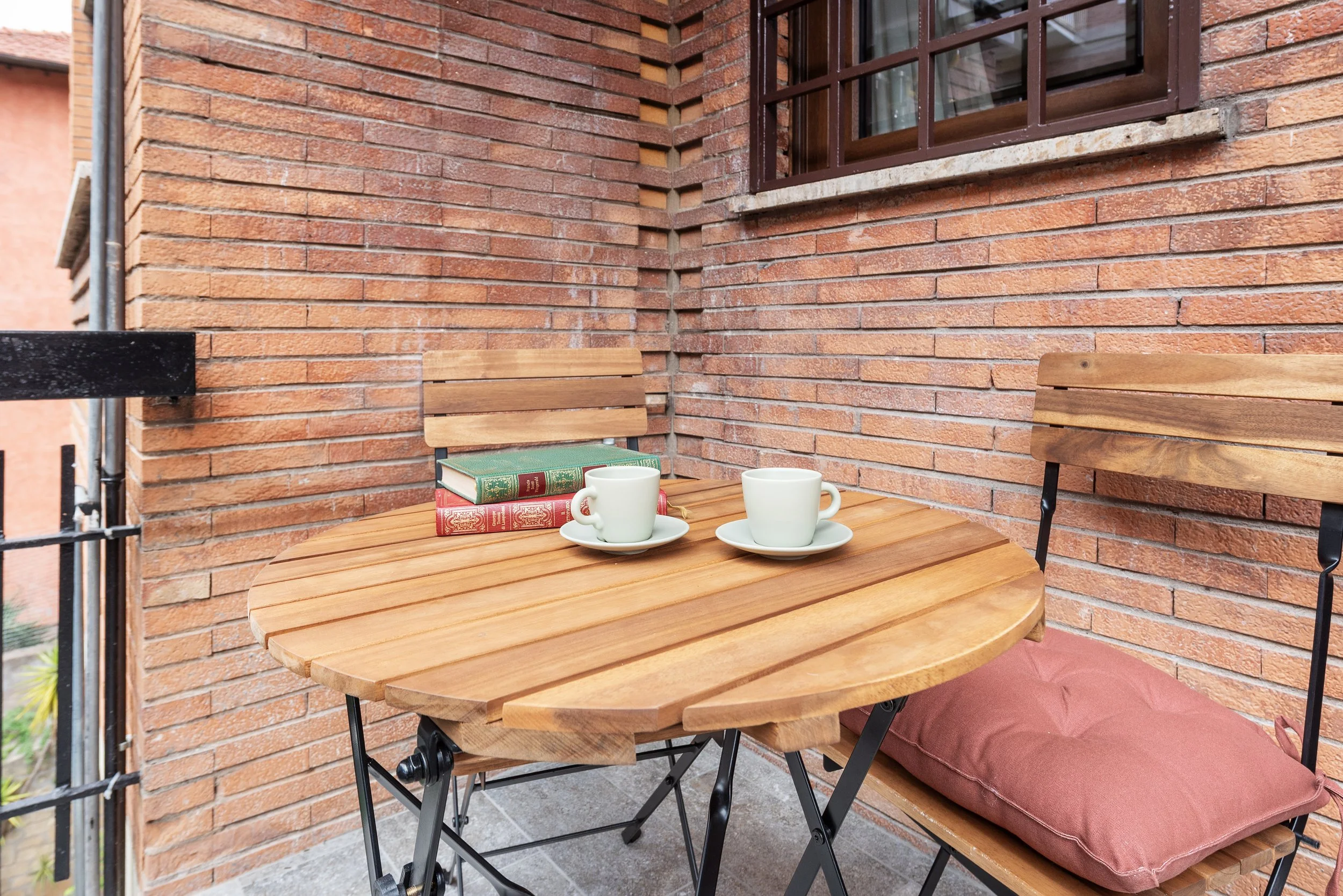 A small round wooden patio table with two white coffee cups on saucers, two hardcover books, and a brown cushion on a wooden chair with a metal frame, set against a brick wall with a window.