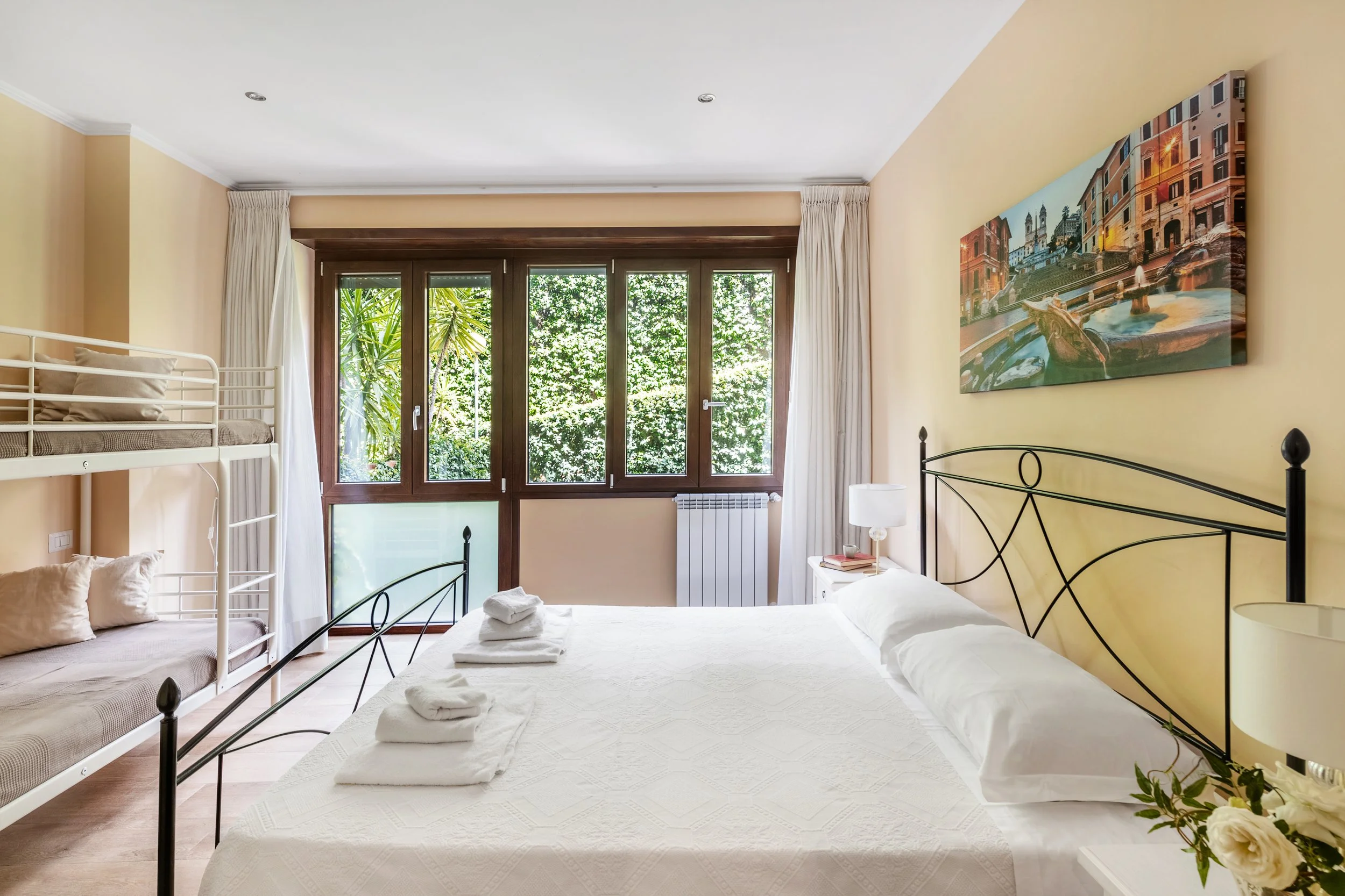 A bedroom with a large bed, a bunk bed on the side, a window with trees outside, and a nightstand with a lamp.