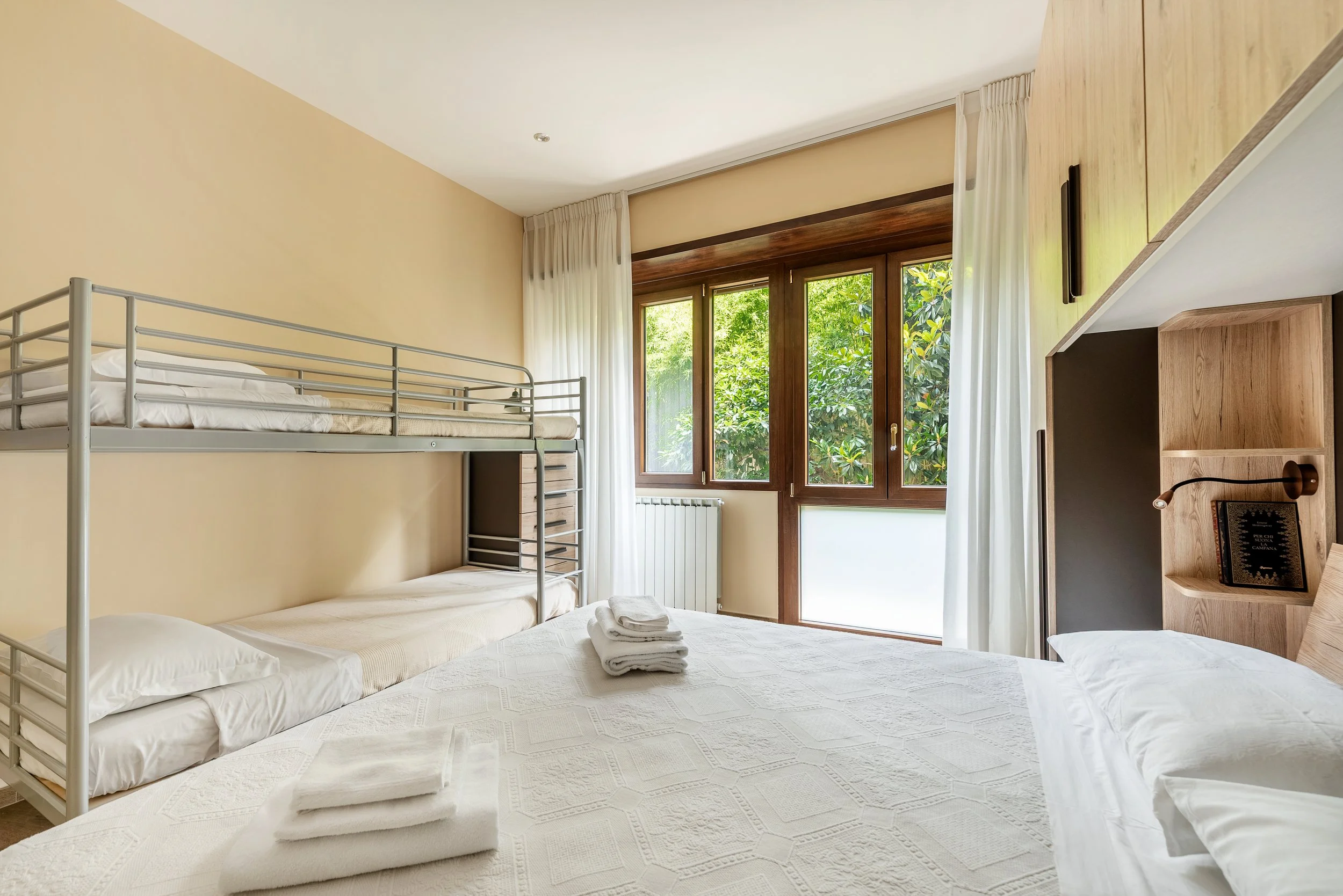 A bright bedroom with a large bed and a set of bunk beds, a window with trees outside, and neutral-colored walls and bedding.