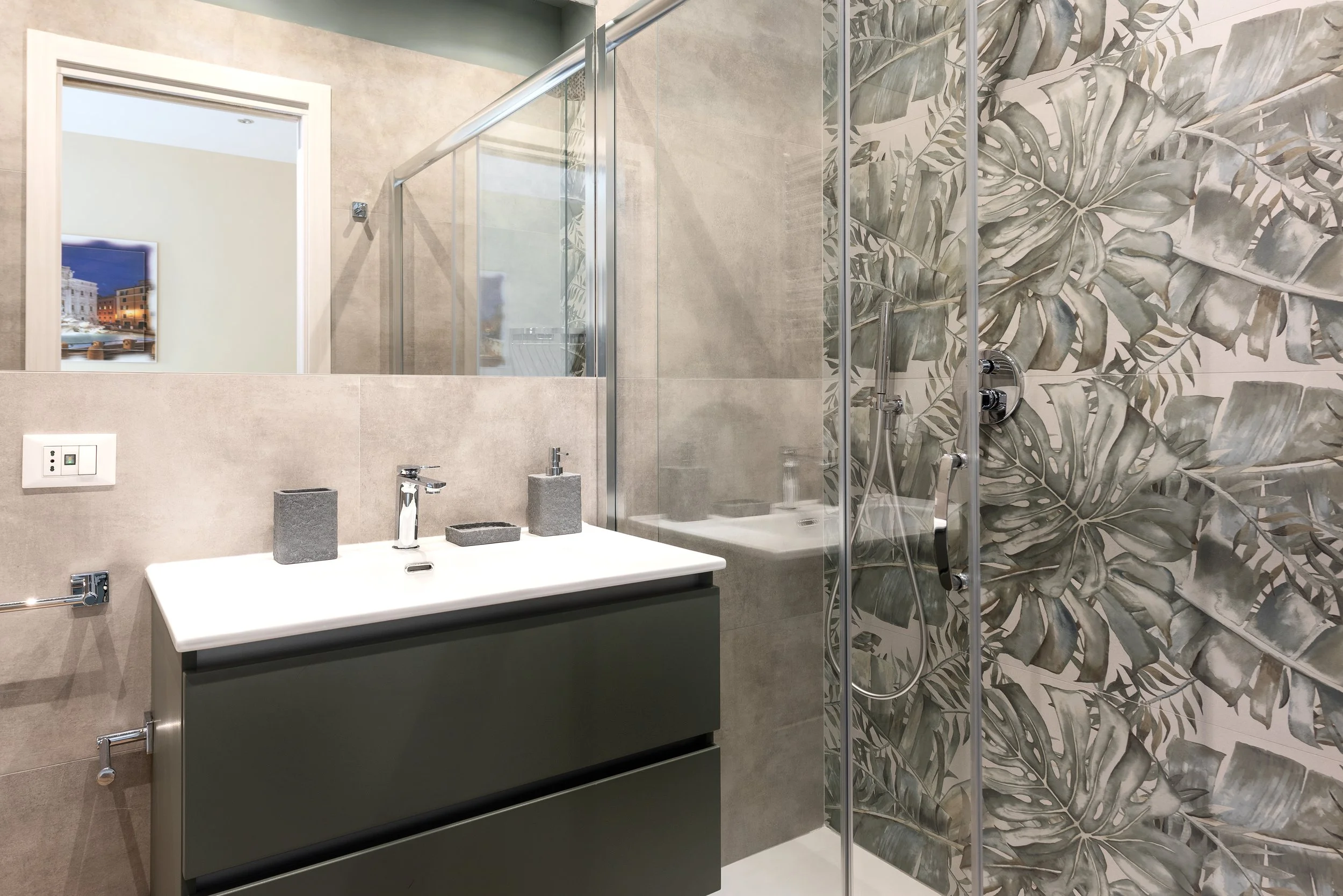 Modern bathroom with beige and botanical tile walls, white sink with gray cabinet, mirror, and glass shower enclosure with leaf-patterned wall.