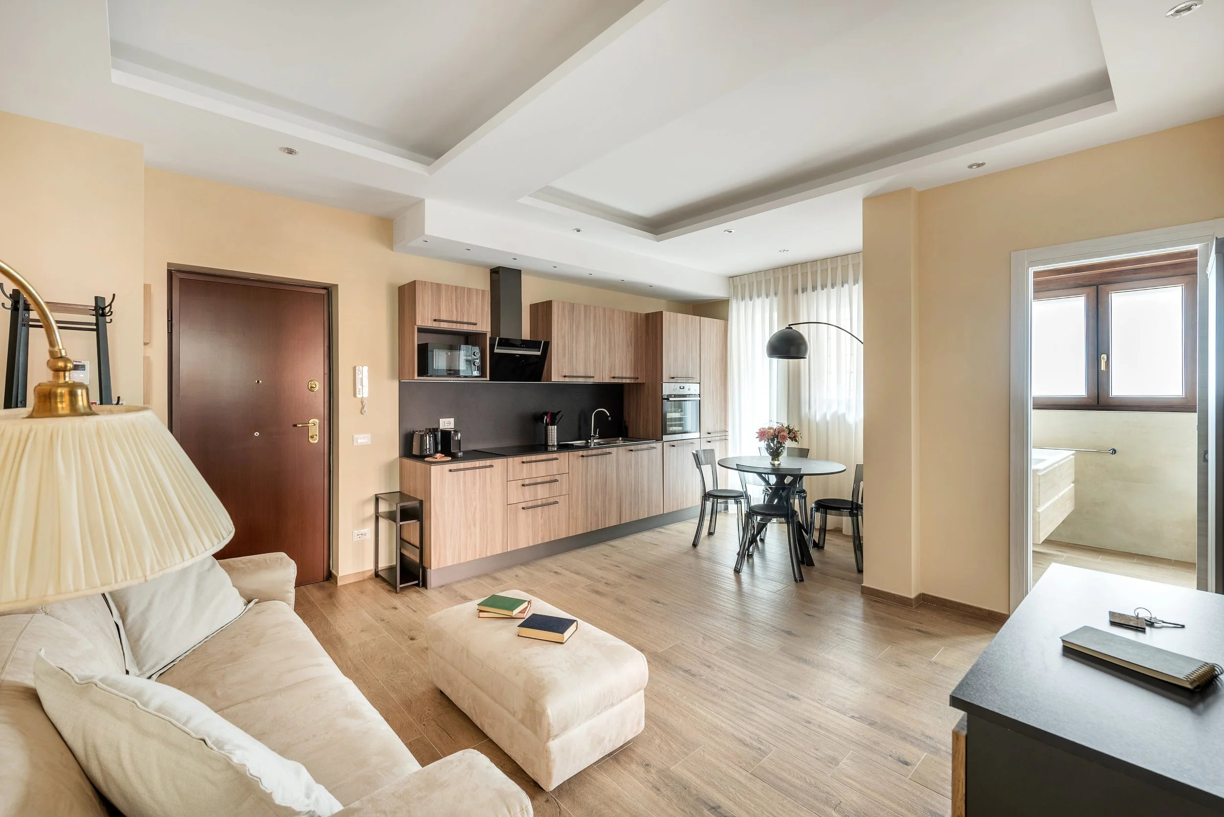 Living room and kitchen with wooden cabinets, round dining table with four chairs, beige sofa with books, and a door leading to a balcony with a bathtub visible outside.