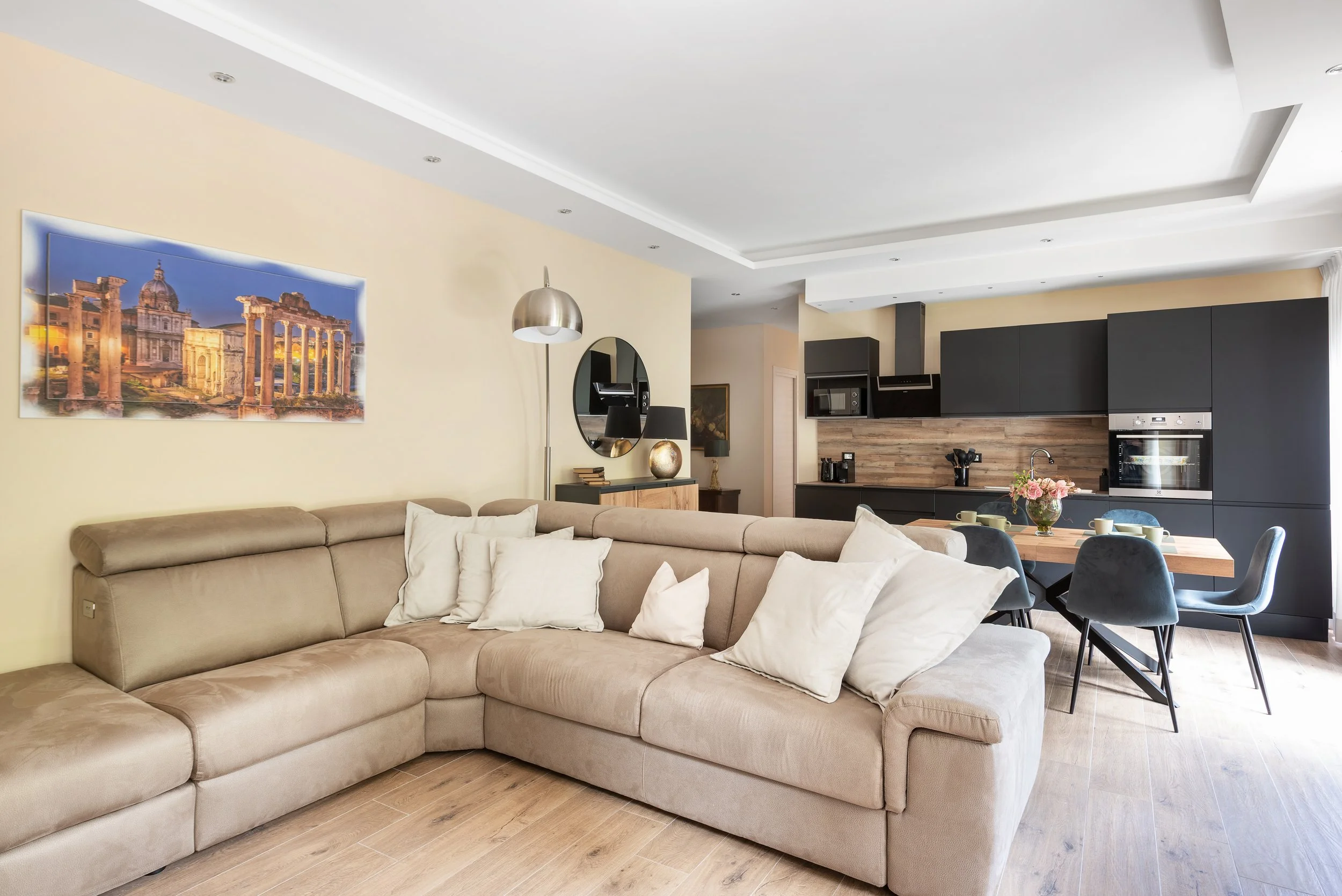 Modern living room and kitchen with beige sectional sofa, wall art, round mirror, floor lamp, and black kitchen cabinets with wooden backsplash, dining table with chairs and floral centerpiece.