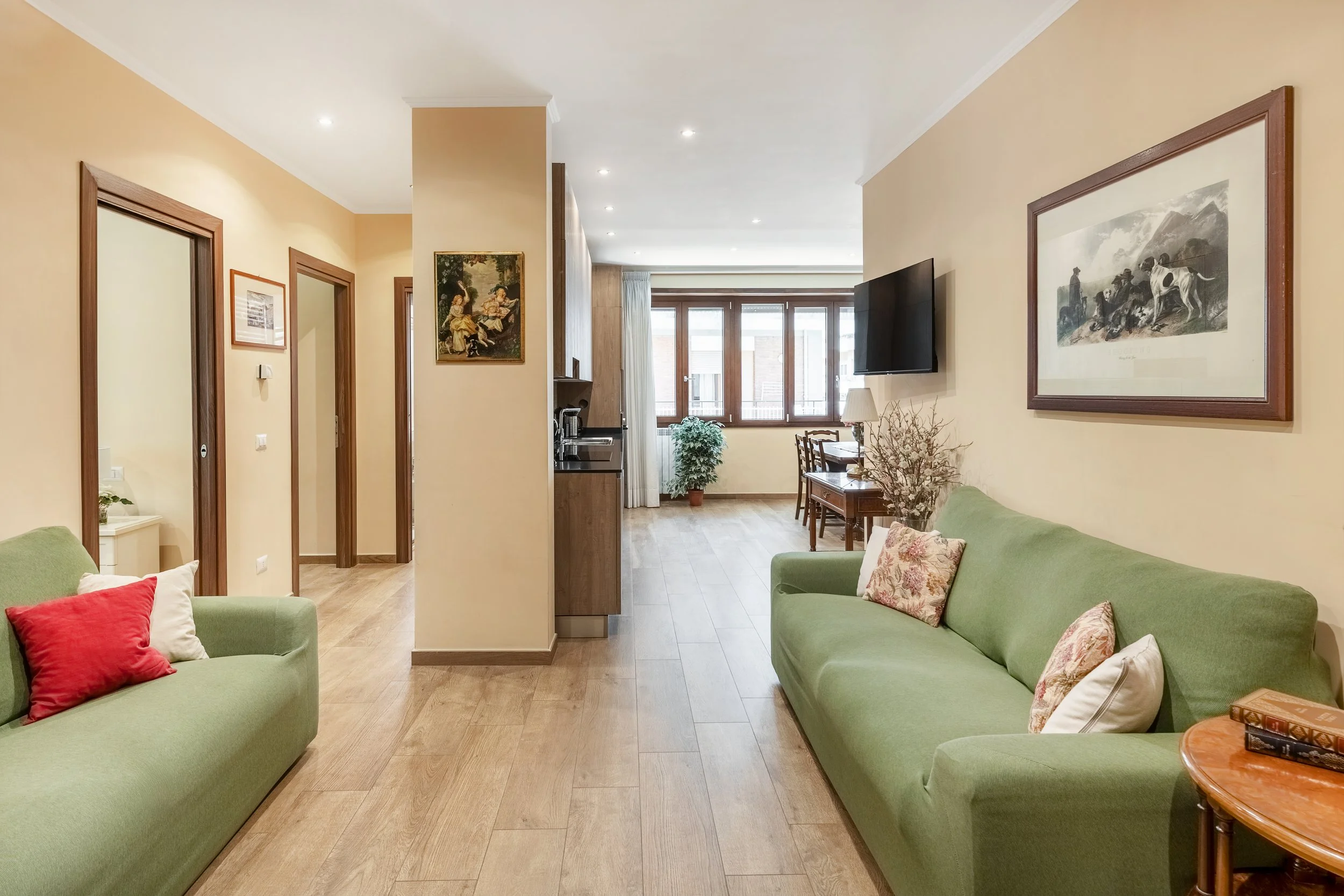 Living room with green sofas, wooden flooring, wall art, a flat-screen TV, and a dining area near large windows.