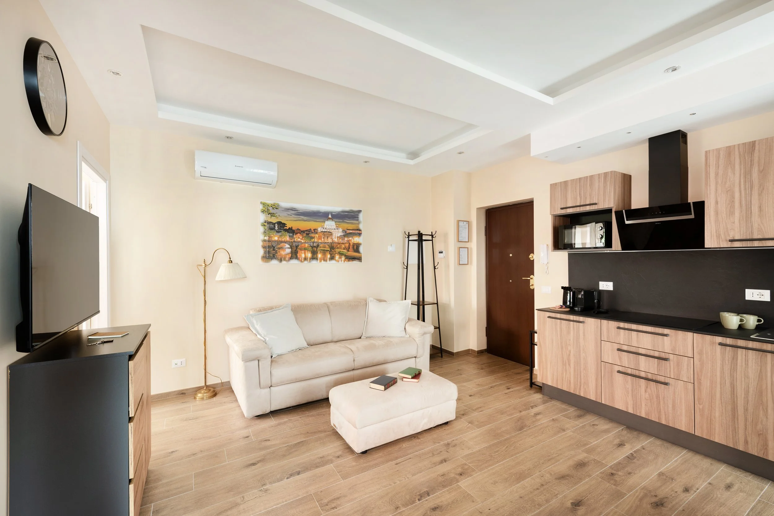 Living room with beige sofa, white pillows, wall art, TV, wooden cabinet, coffee table, and kitchen area with wooden cabinets and black countertops.