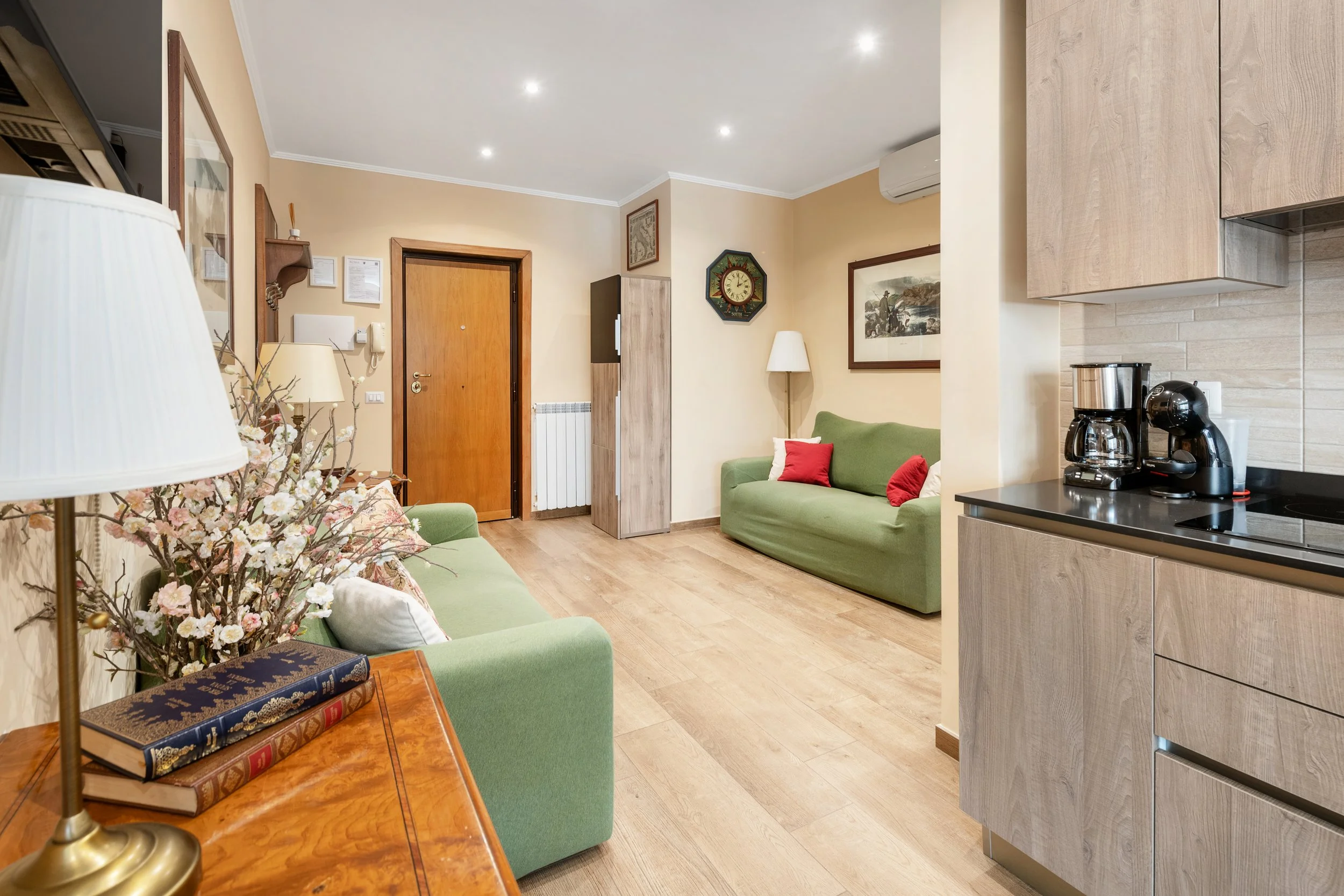 Living room with green sofas, wooden floor, cream walls, decorative paintings, an entrance door, a small kitchenette with coffee maker, and various furniture and decorations.