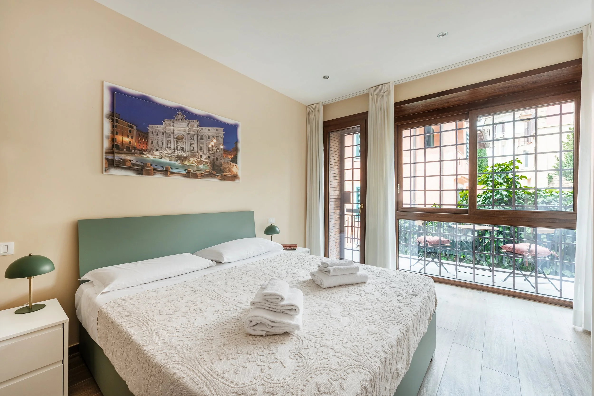 A bedroom with a large bed, green headboard, white bedding, and four folded towels on top. There are two small green bedside lamps, a nightstand, and a window with white curtains and a view of a balcony with chairs and greenery outside.