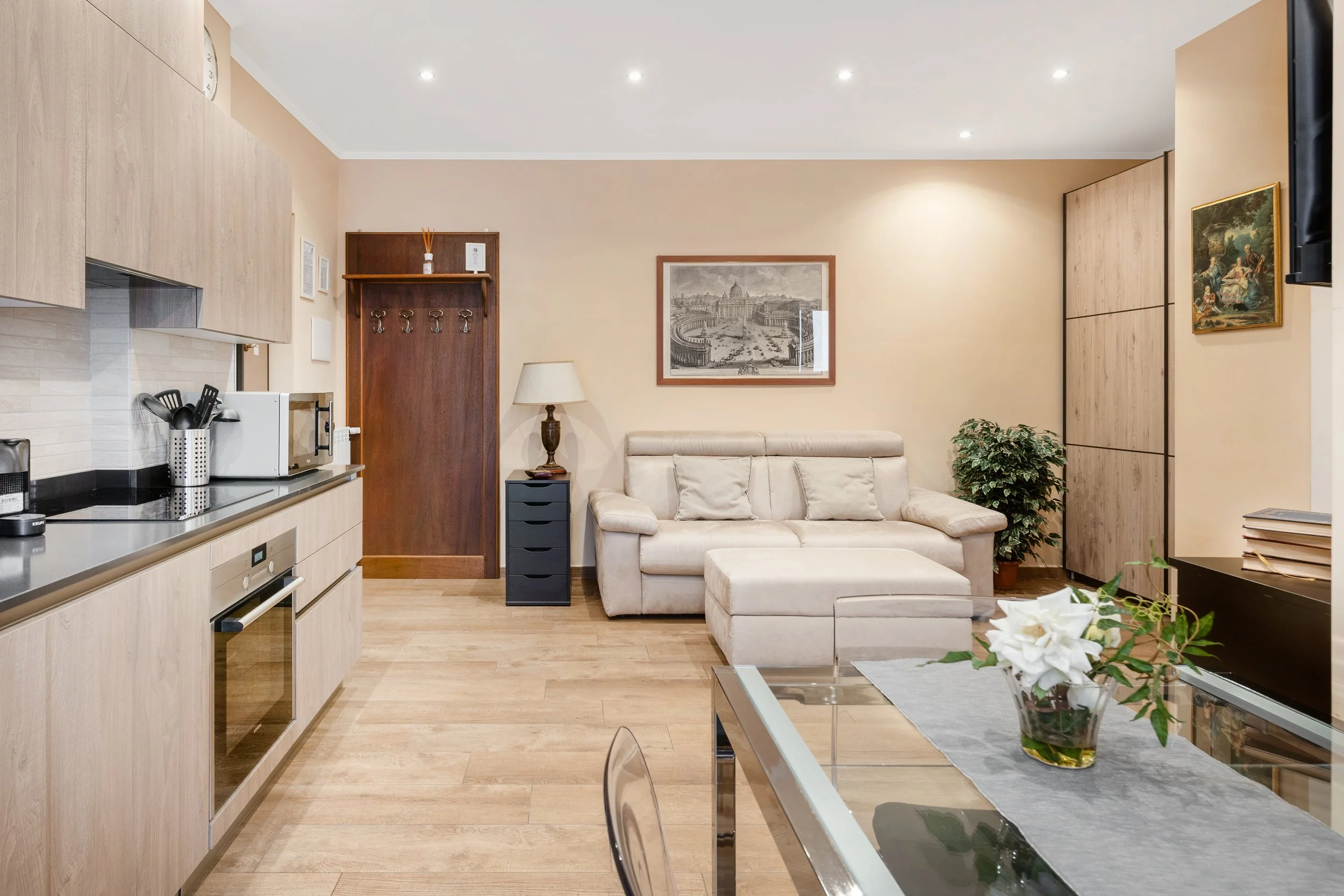 Living room with a beige sectional sofa, a wooden side table with a lamp, framed artwork on the wall, a potted plant, and a modern kitchen area with light wood cabinets and stainless steel appliances.