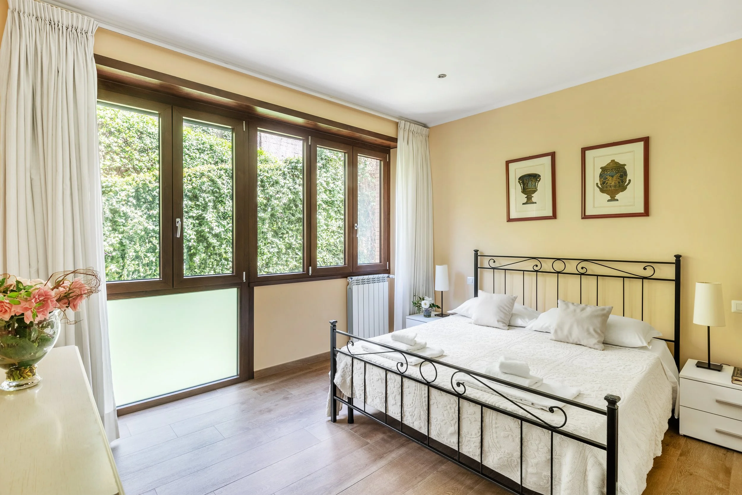 Bright hotel bedroom with a large bed, white bedding, and pillows. There are two nightstands with lamps and a vase of flowers, framed artwork above the bed, large windows with frosted and clear glass, curtains, and a wood floor.
