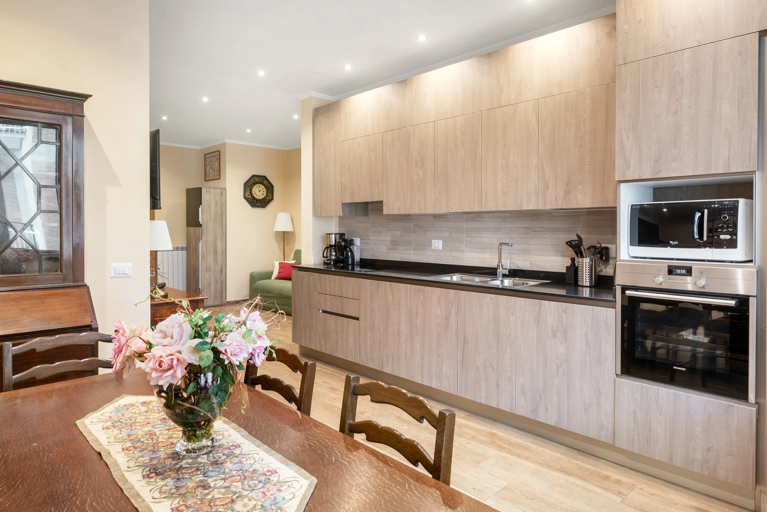 Interior view of a kitchen with a wooden dining table and a vase of pink flowers, modern cabinets, built-in oven and microwave, coffee maker, sink, and a living room area with green sofa and lamps in the background.