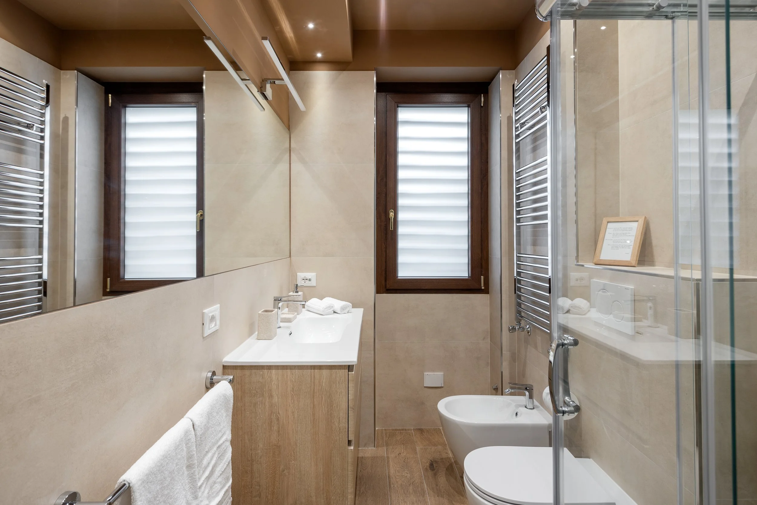 Modern bathroom with beige tiles, wooden accents, two windows with frosted glass, a white sink with toiletries, a towel rack with a towel, a bidet, and a glass shower enclosure.