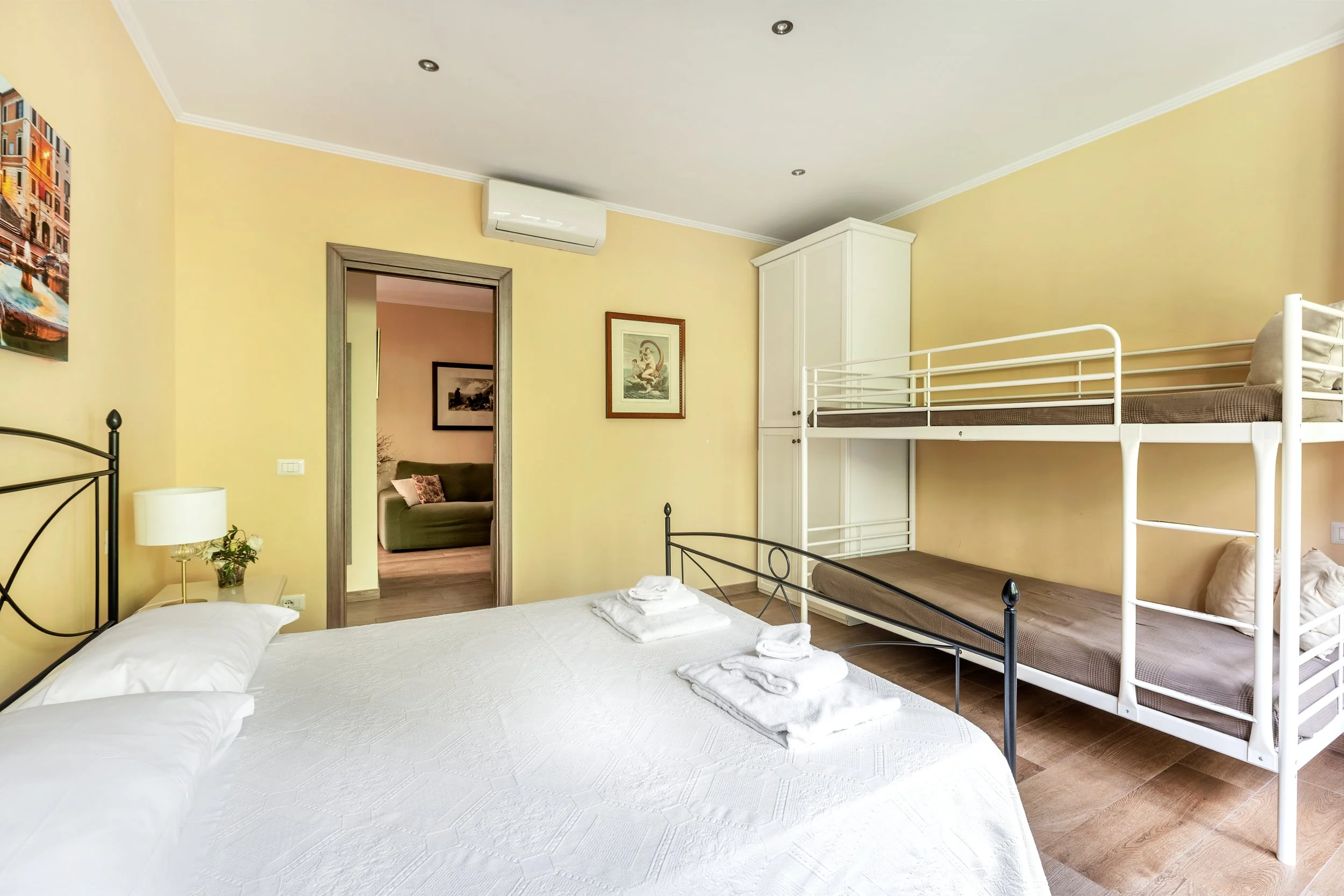 Bedroom with a double bed, folded towels, a nightstand with a lamp and flowers, and a bunk bed with two mattresses, yellow walls, framed artwork, and an air conditioner above the door leading to another room with a sofa.