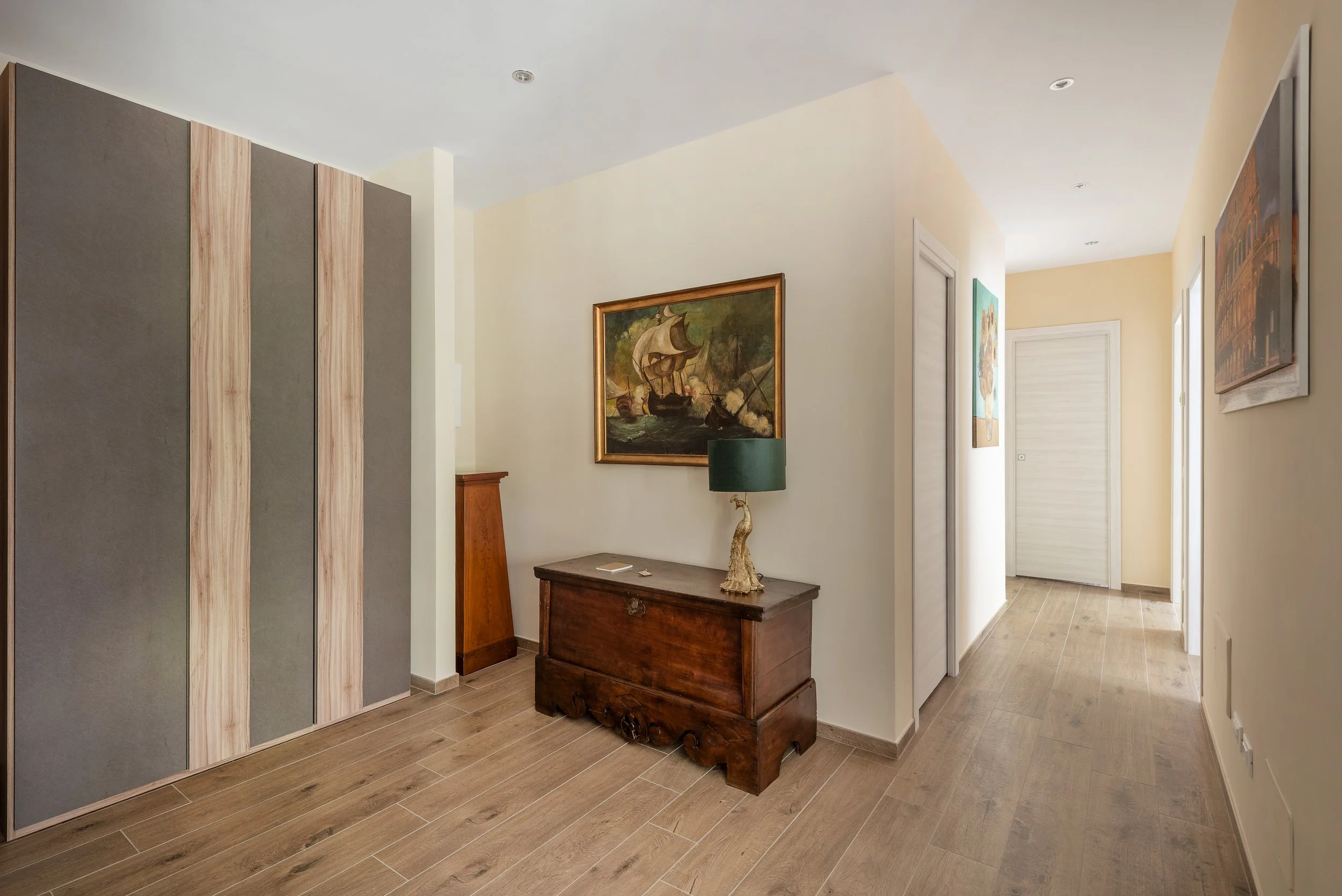 Interior of a hallway with wooden flooring, a wooden dresser with a decorative lamp, framed paintings on the walls, and a large built-in wardrobe on the left side.