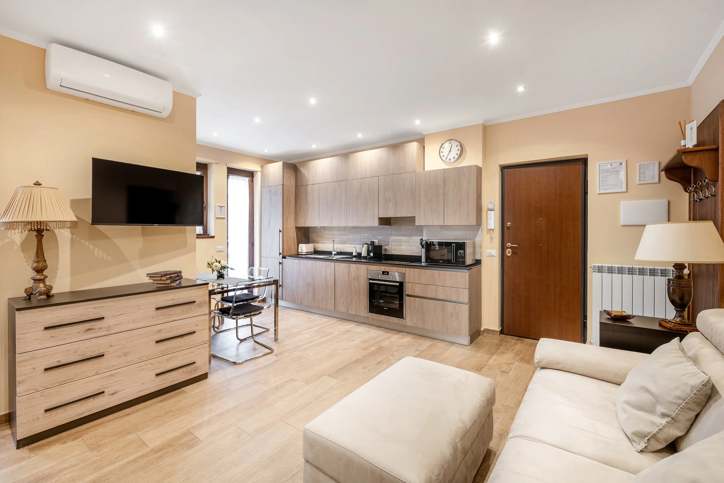 Modern living room with beige walls and wooden floors, featuring a wall-mounted flat-screen TV, a wooden sideboard with a lamp and decorative items, a beige sofa, a small dining table with chairs, and a kitchenette with light wood cabinets and applia